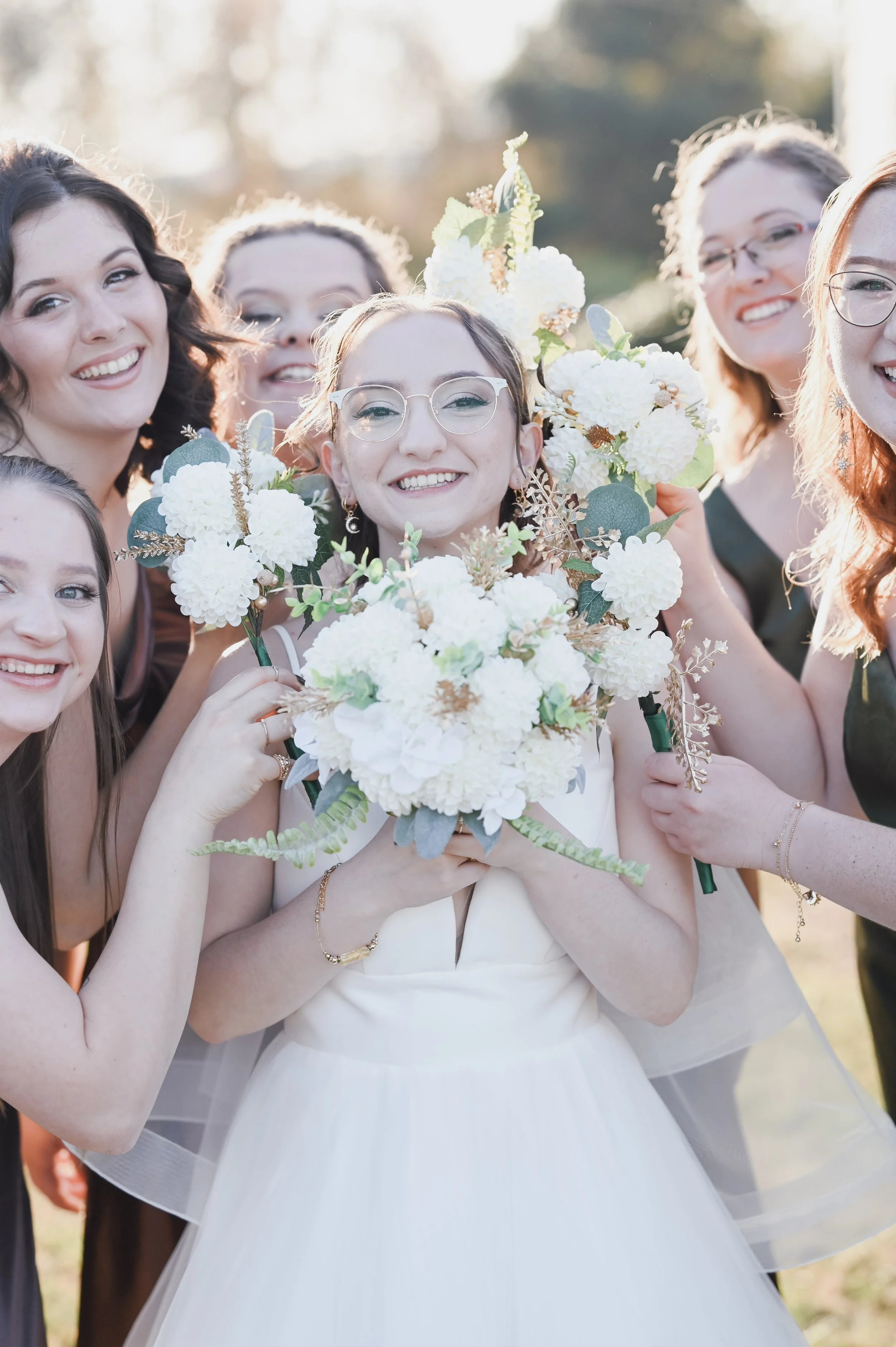 A bride holding a bouquet of white flowers surrounded by her bridesmaids at a wedding celebration outdoors.