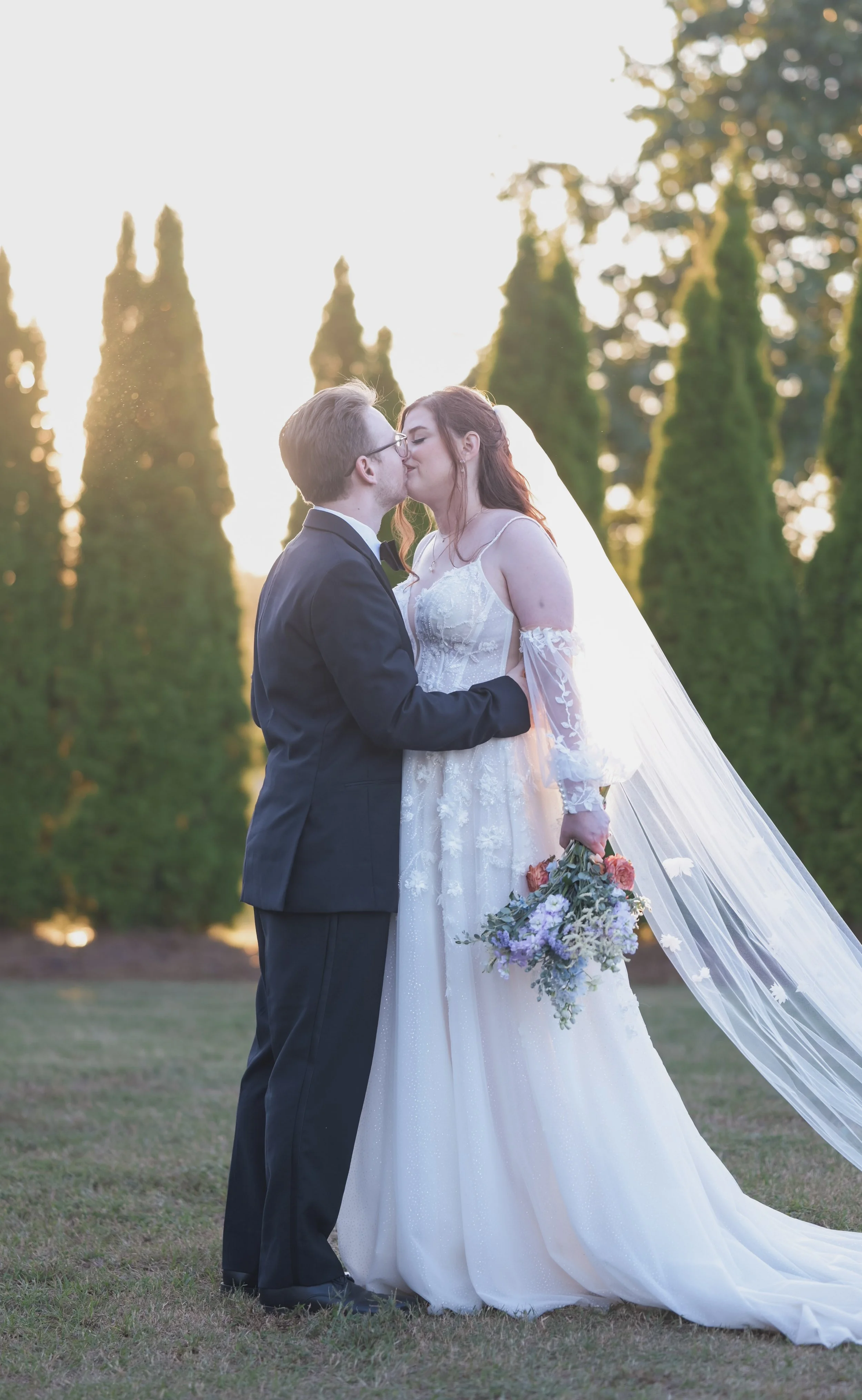 A bride and groom kissing outdoors during sunset, with tall green trees in the background. The bride is holding a colorful bouquet and wearing a white wedding dress with long lace sleeves, while the groom is dressed in a black tuxedo.