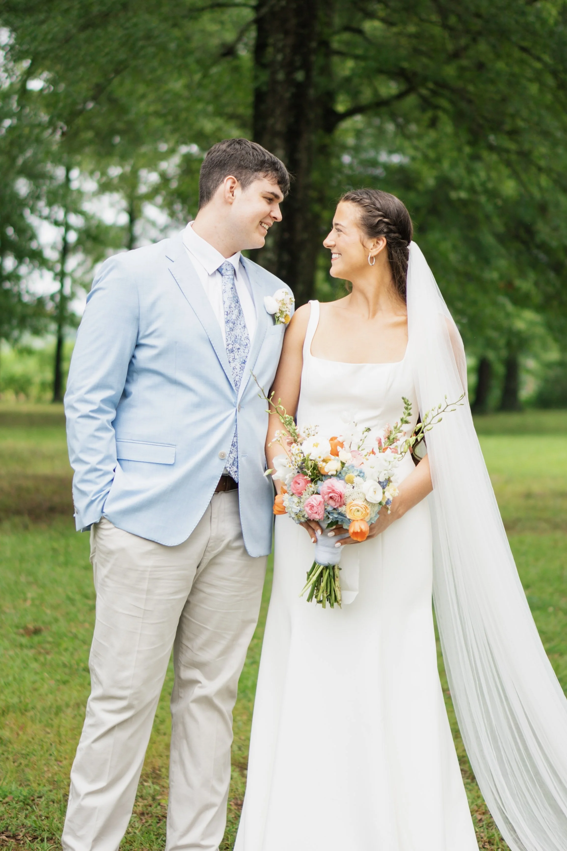 A bride and groom happy in an outdoor setting, with the groom wearing a light blue suit and the bride in a white gown holding a colorful bouquet, standing on grass with trees in the background.