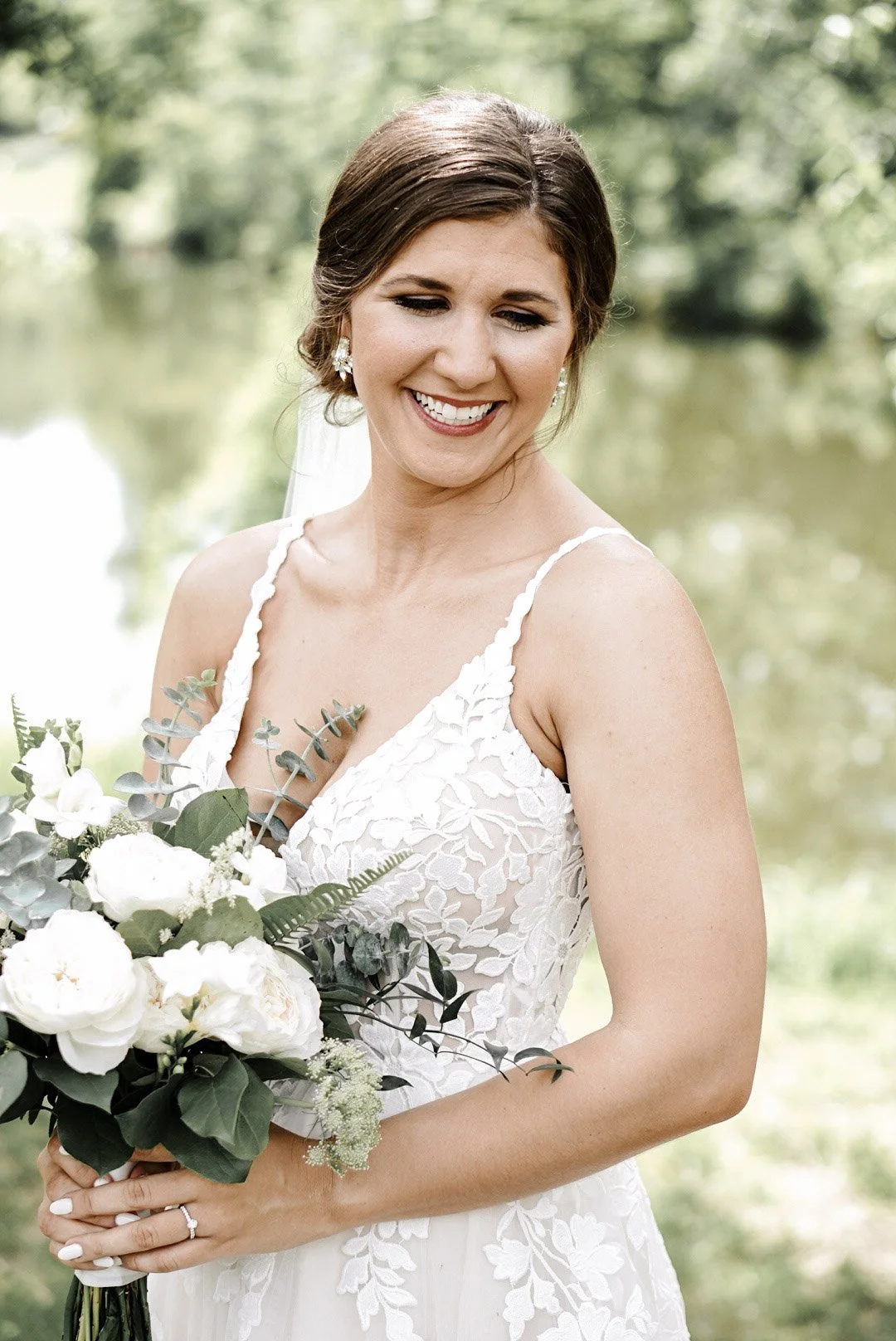 A smiling bride with brown hair styled in an updo, wearing a white lace wedding dress with thin straps, holding a bouquet of white flowers and greenery, outdoors near a body of water with trees in the background.