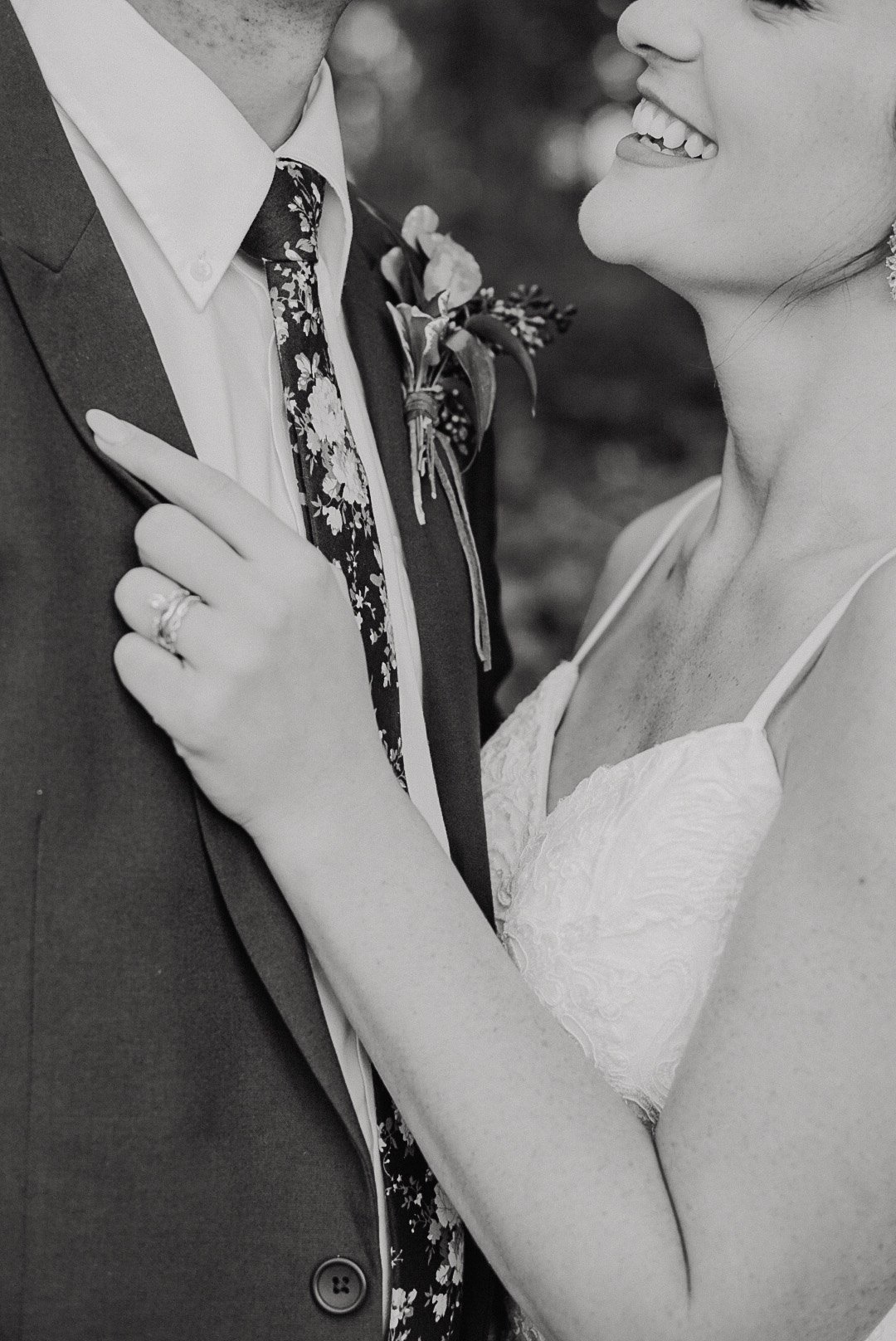 A black and white photograph of a smiling bride and groom close together, showing the bride's hand with a wedding ring on her finger, touching the groom's chest and pointing at his boutonniere.