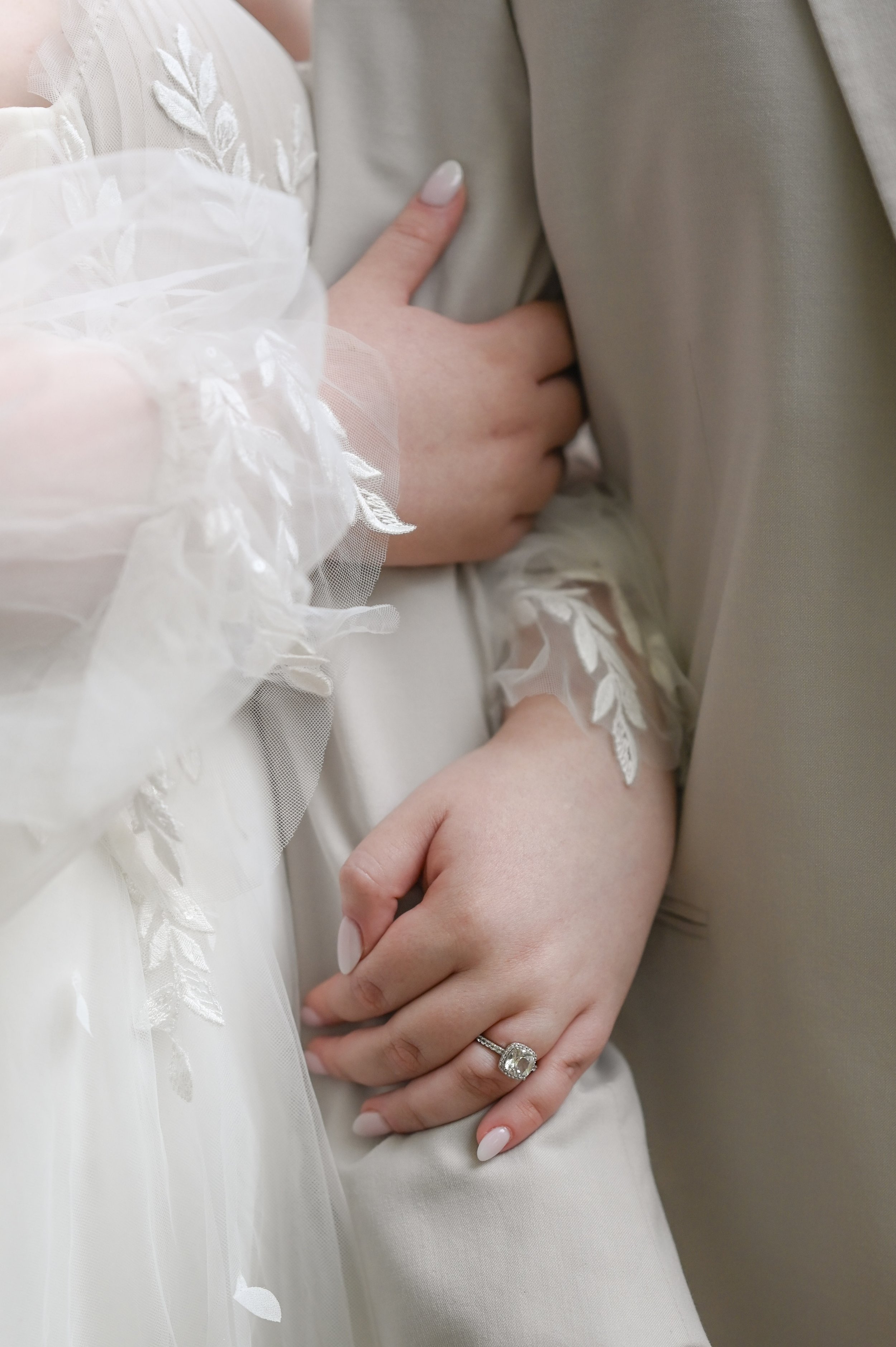 Close-up of a woman's hand with an engagement ring, resting on a man’s arm, both wearing elegant formal attire, with lace and embroidered details.