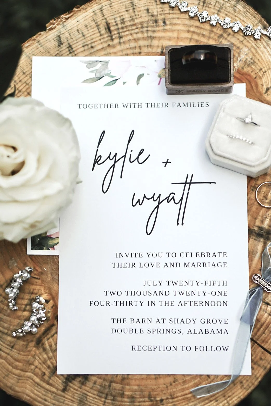 Wedding invitation with the names Kylie and Wyatt, surrounded by wedding accessories including rings, a flower, earrings, and rings in a box on a wooden surface.