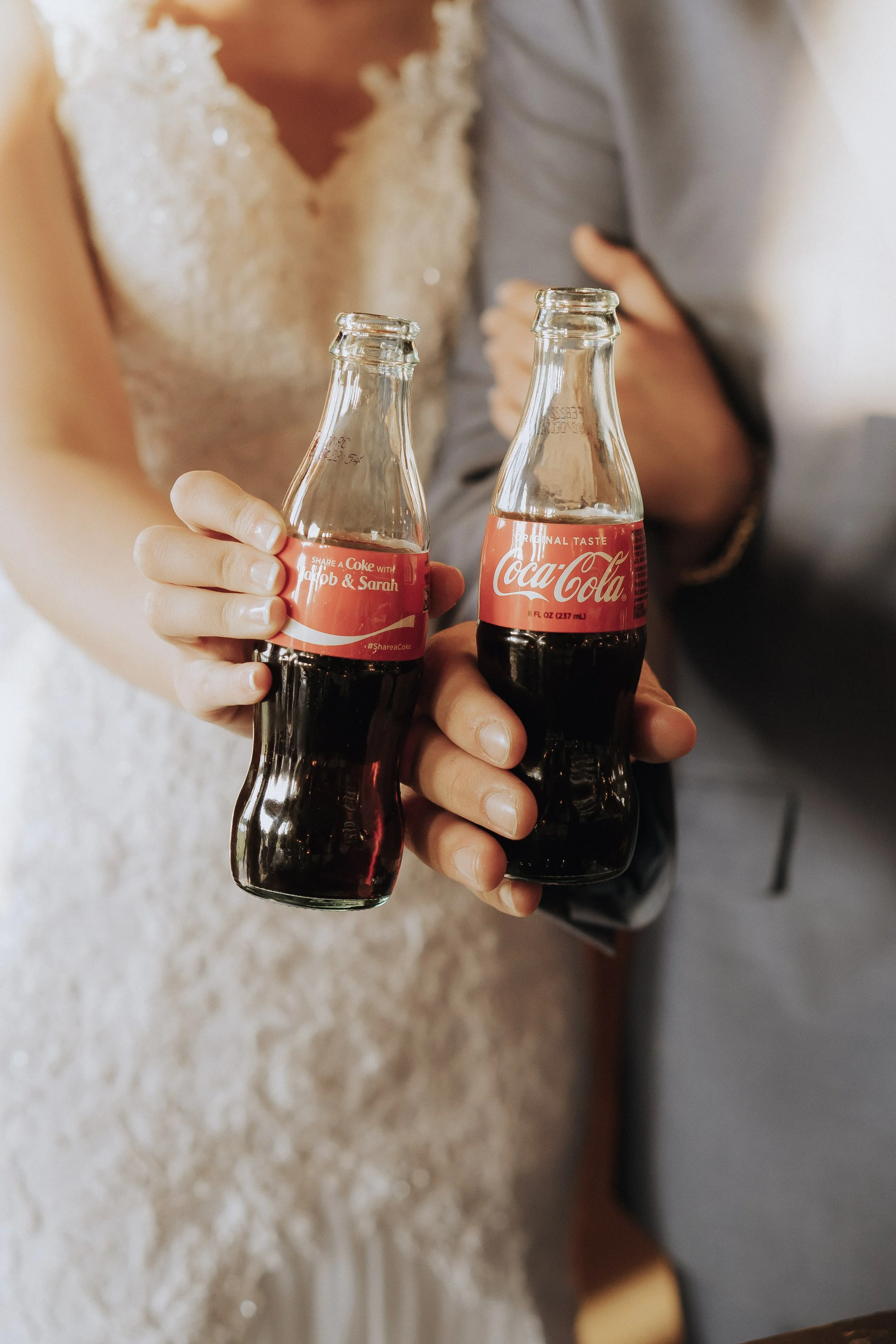 Close-up of two hands holding glass Coca-Cola bottles, one with a personalized label, at a wedding celebration.