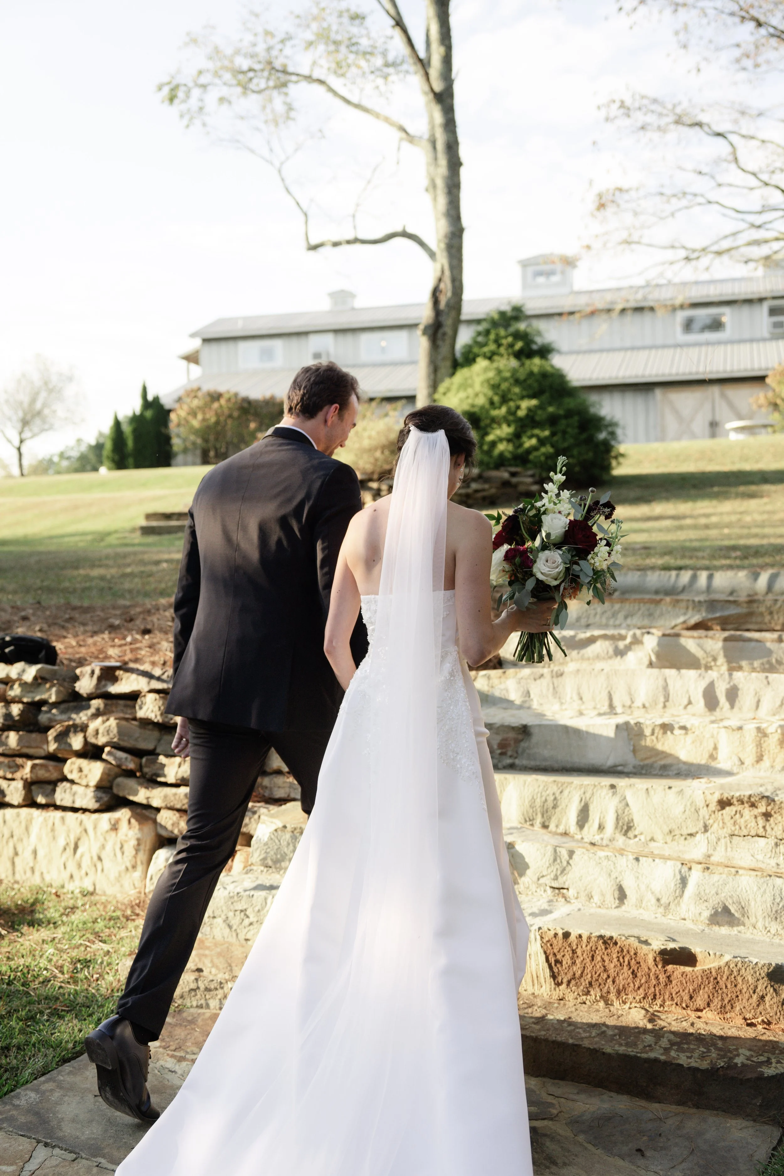 Bride and groom ascending stone steps outdoors, bride holding a bouquet of white and dark red flowers, groom wearing a black suit, in a natural setting with trees and a large building in the background.