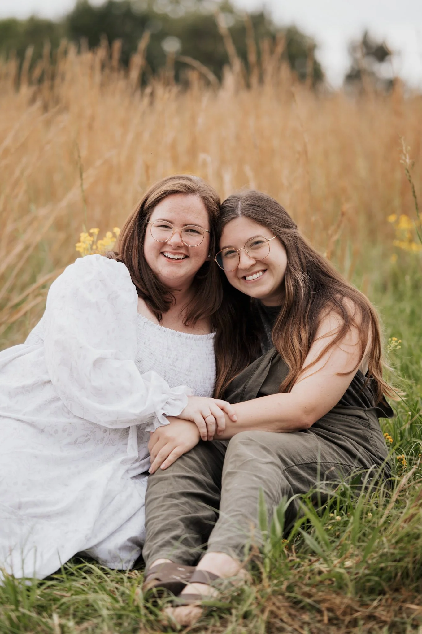 Two women sitting on the grass in a field with tall golden grass, smiling and holding hands, wearing glasses and casual clothing.