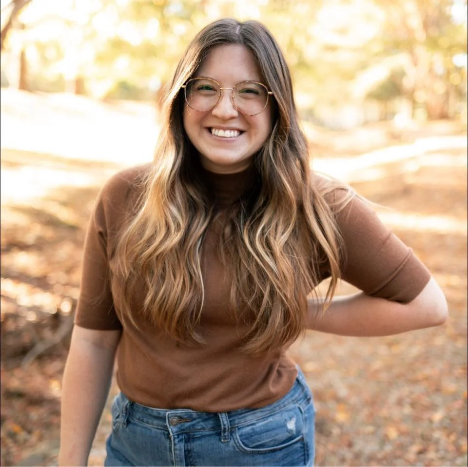 A young woman with long, wavy hair and glasses, smiling outdoors in a wooded area during autumn.