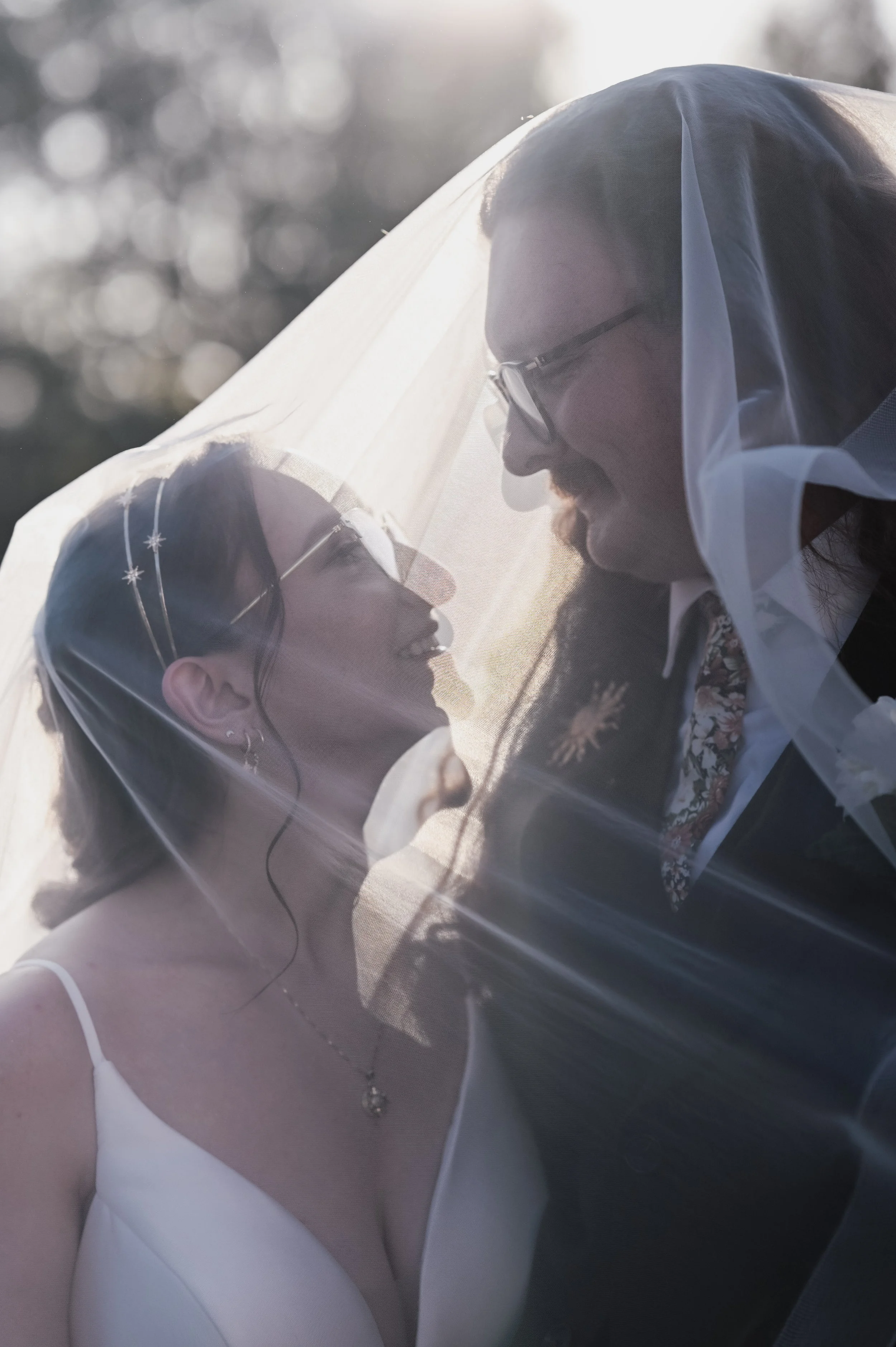 A bride and groom face each other under a sheer veil during a wedding, with sunlight behind them creating a warm glow.