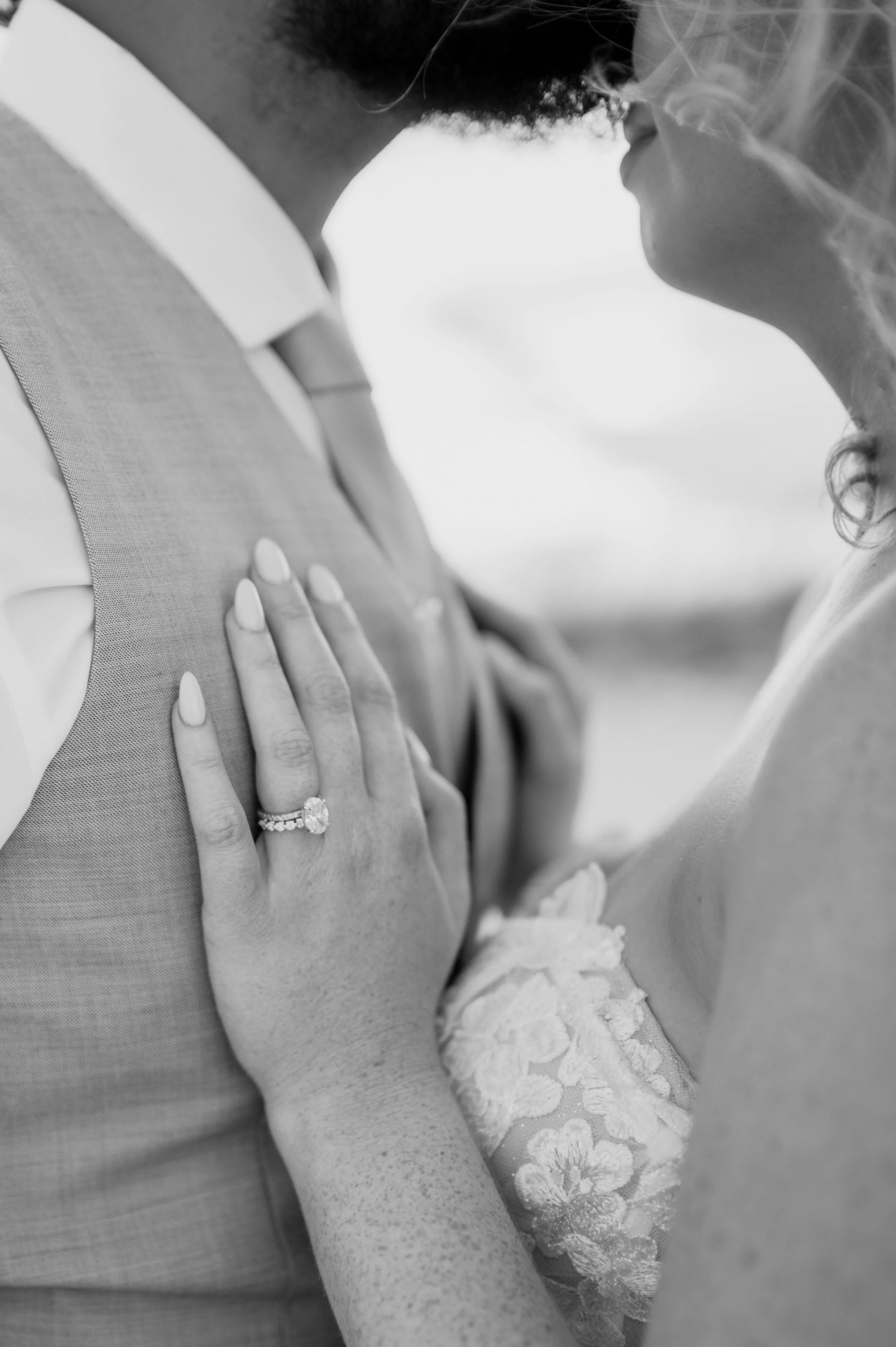Close-up of a bride and groom in an intimate moment, with the bride's hand showing an engagement ring resting on the groom's chest. The bride's dress features lace details, and the groom is wearing a vest and tie.