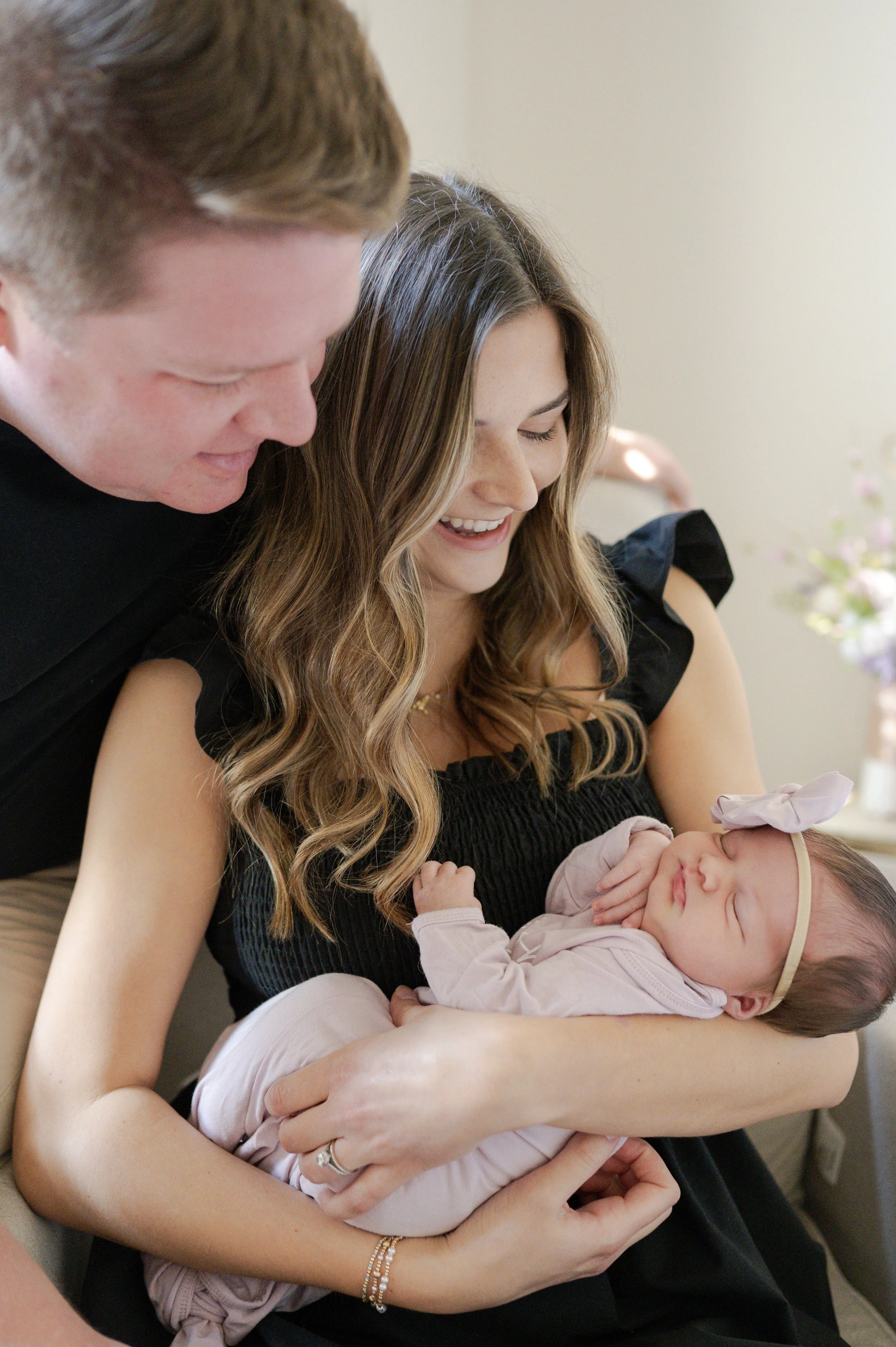Woman holding sleeping baby girl with bow headband, man looking at her, in a cozy indoor setting.