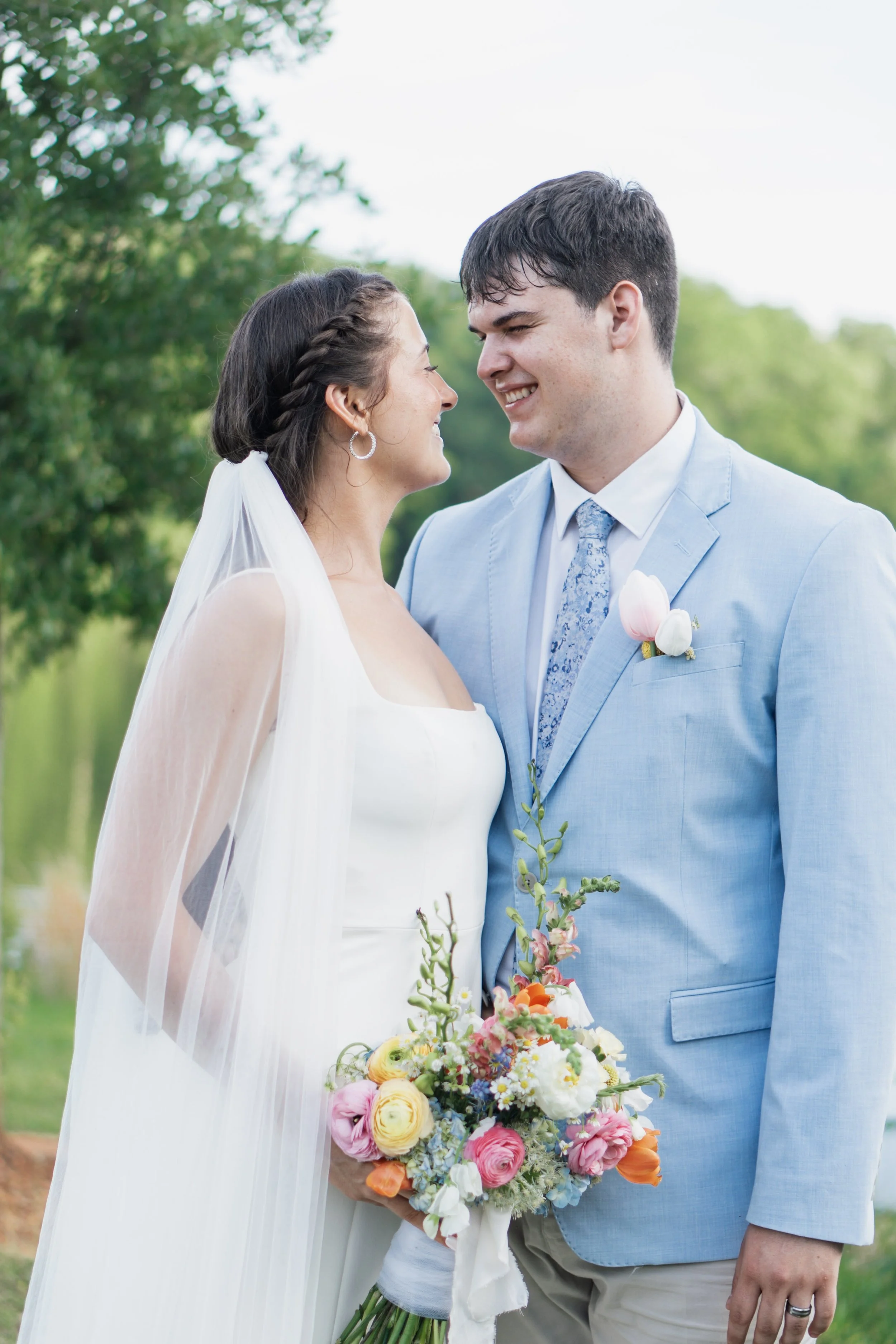 A bride and groom standing close together outdoors, smiling at each other with a background of green trees. The bride is holding a colorful bouquet of flowers, and the groom is wearing a light blue suit with a boutonnière.