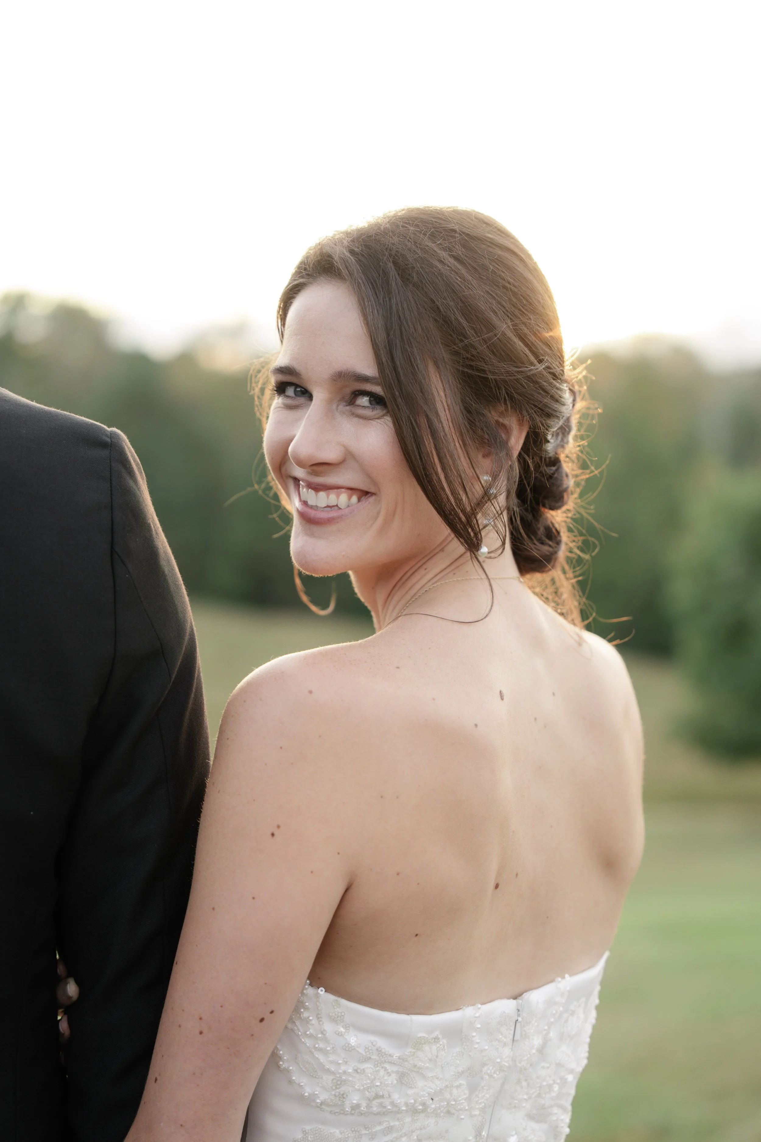 A smiling bride with dark brown hair styled in an updo outdoor during sunset, wearing a strapless wedding dress with lace details.