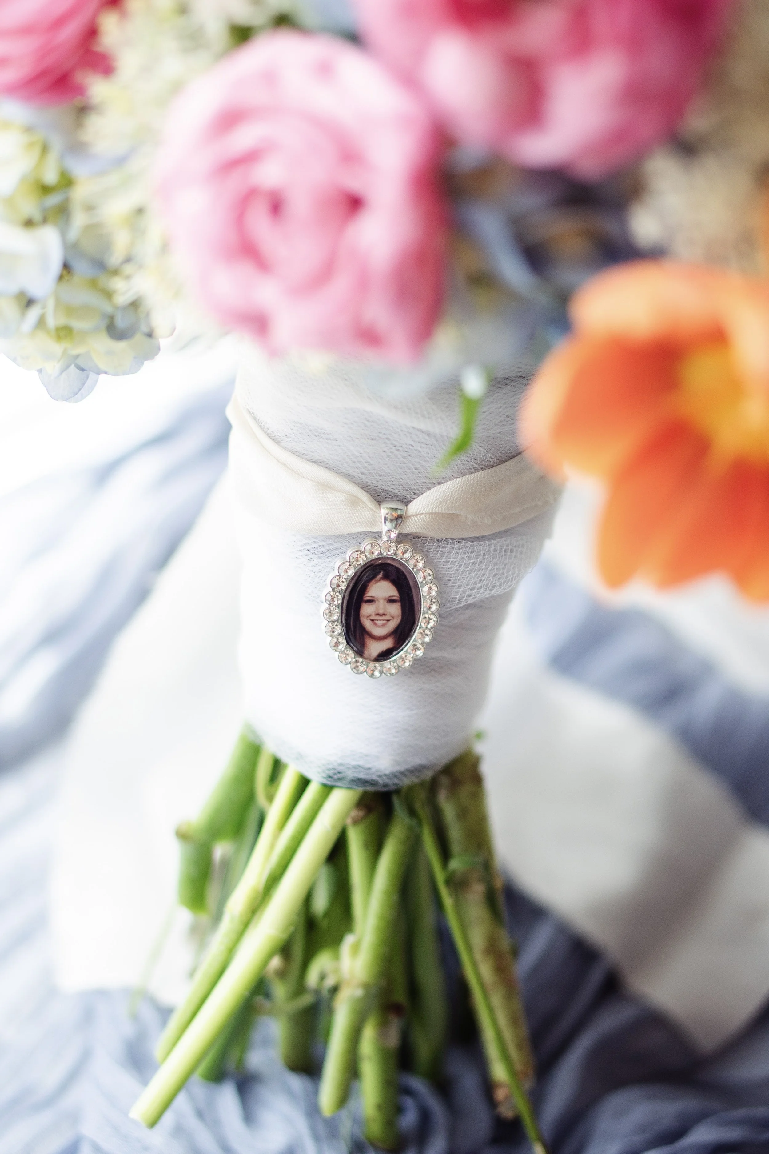 Bouquet of pink, orange, and green flowers wrapped in white ribbon with a photo necklace of a smiling woman attached to it.