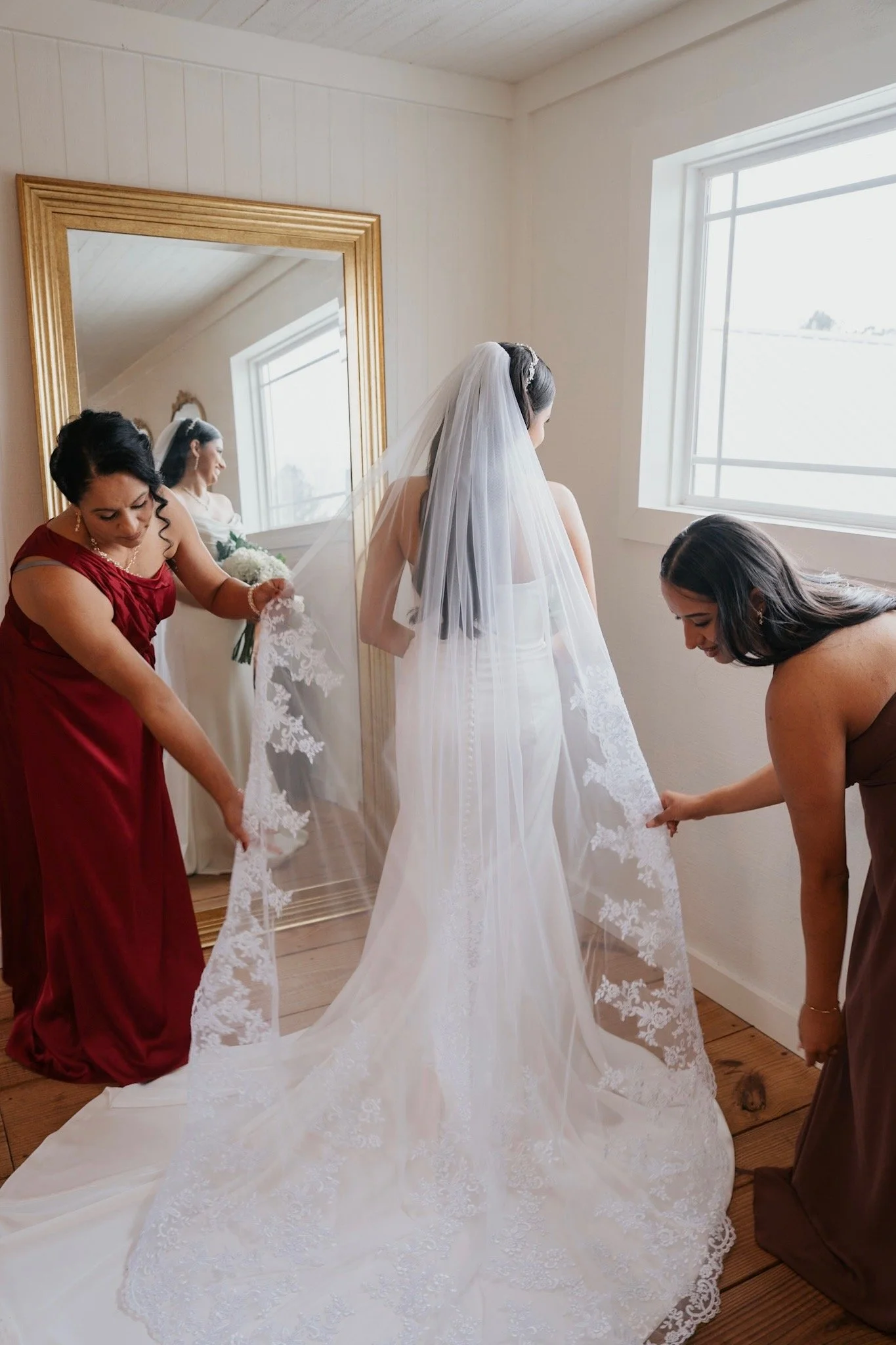 A bride in a white wedding dress with a long veil is being assisted by two women in a room with a large mirror and a window, as they prepare her wedding dress.