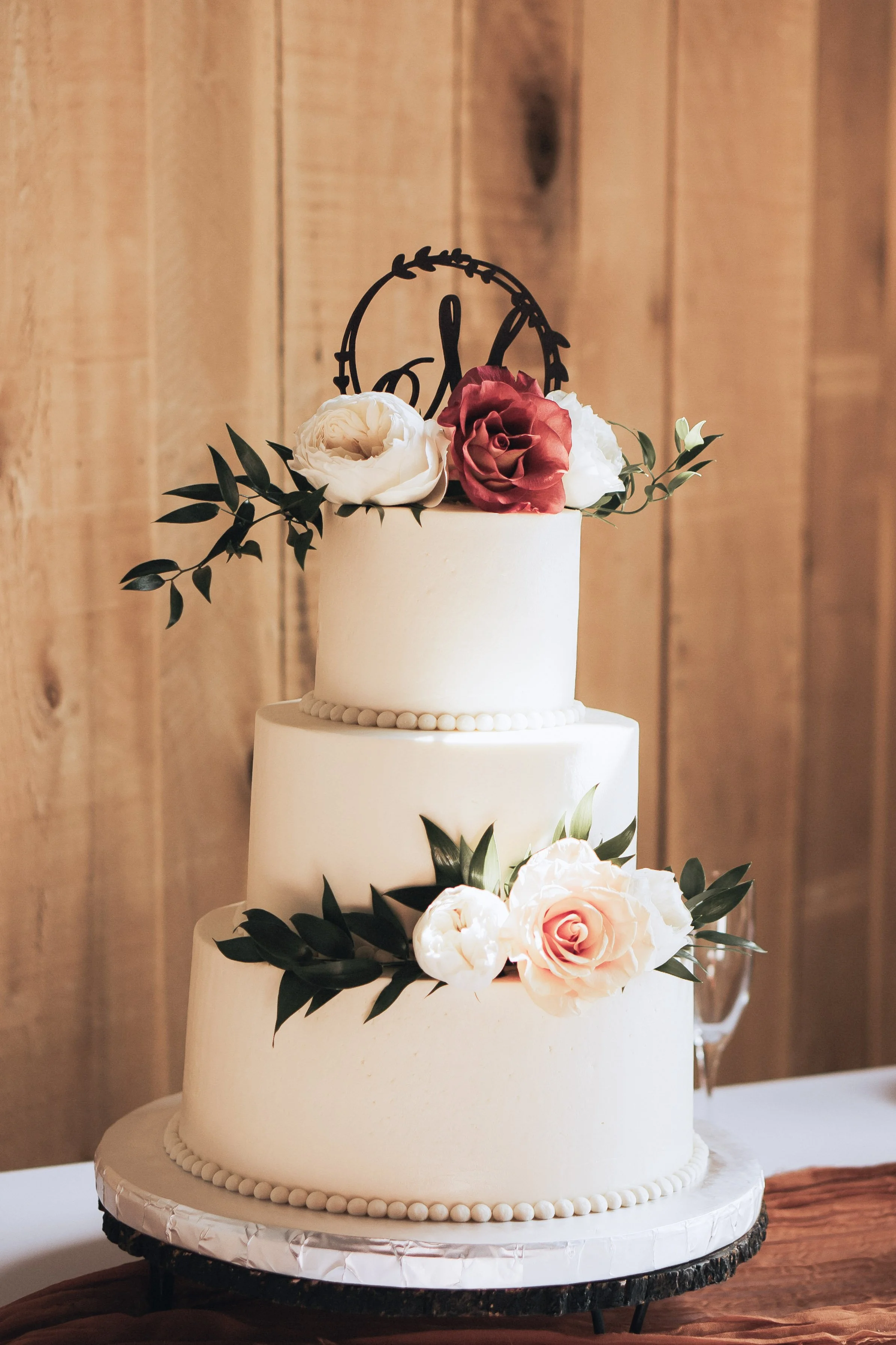 Three-tier white wedding cake decorated with white and pink roses, greenery, and a black cake topper with calligraphy, set against a wooden background.