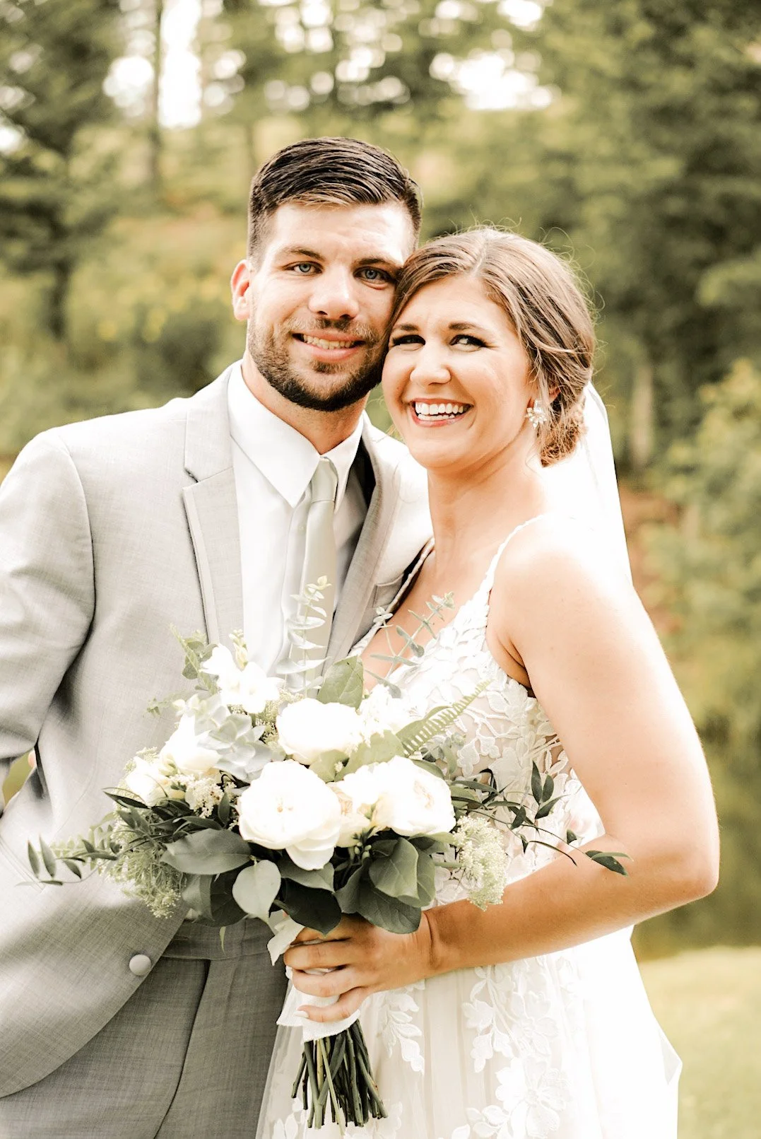 A newlywed couple dressed in wedding attire outdoors. The groom in a light grey suit and tie, and the bride in a white lace wedding dress, holding a bouquet of white roses and greenery. They are smiling and close together with trees in the background