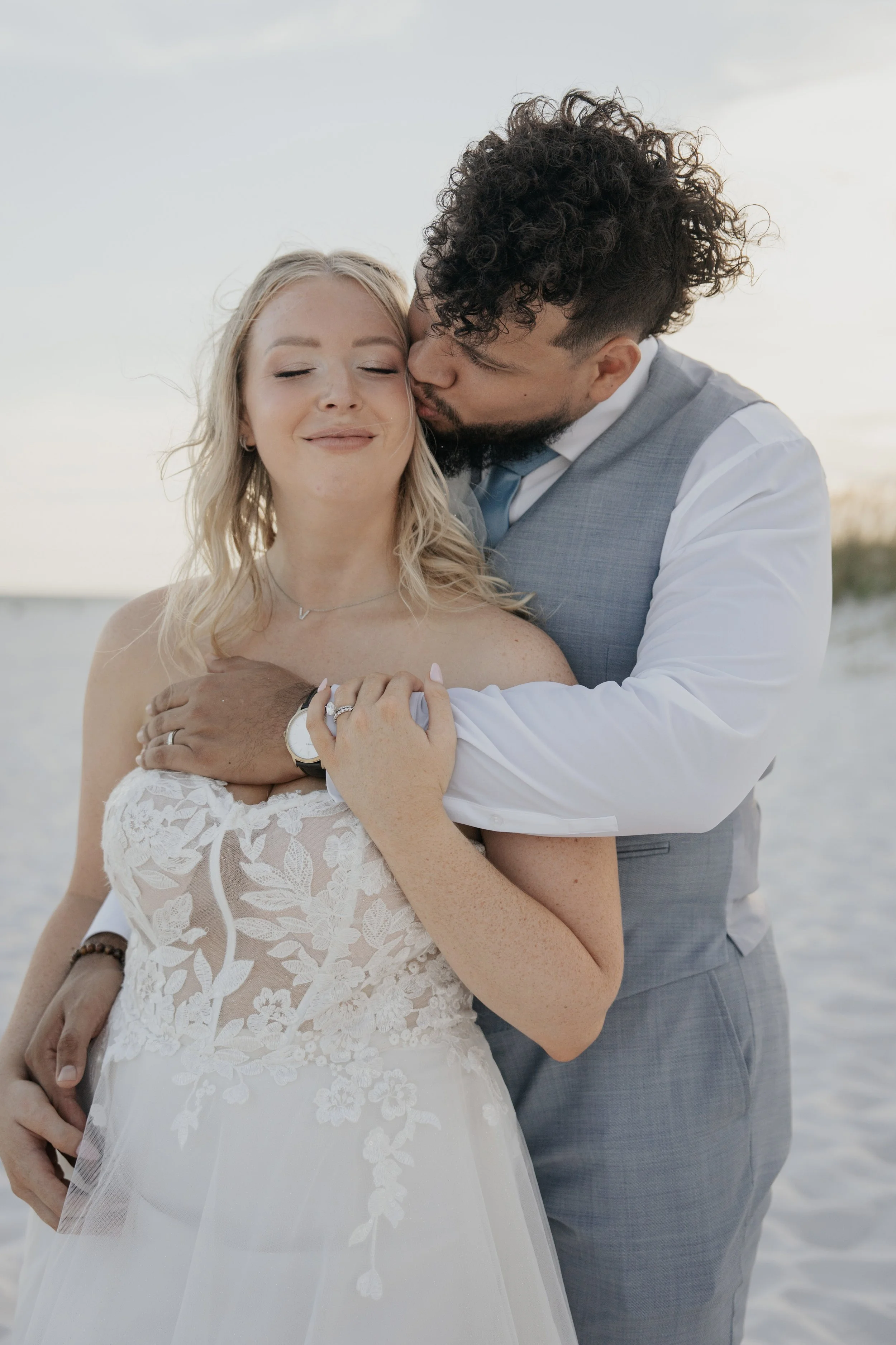 A couple in wedding attire embracing at the beach during sunset, with waves in the background.