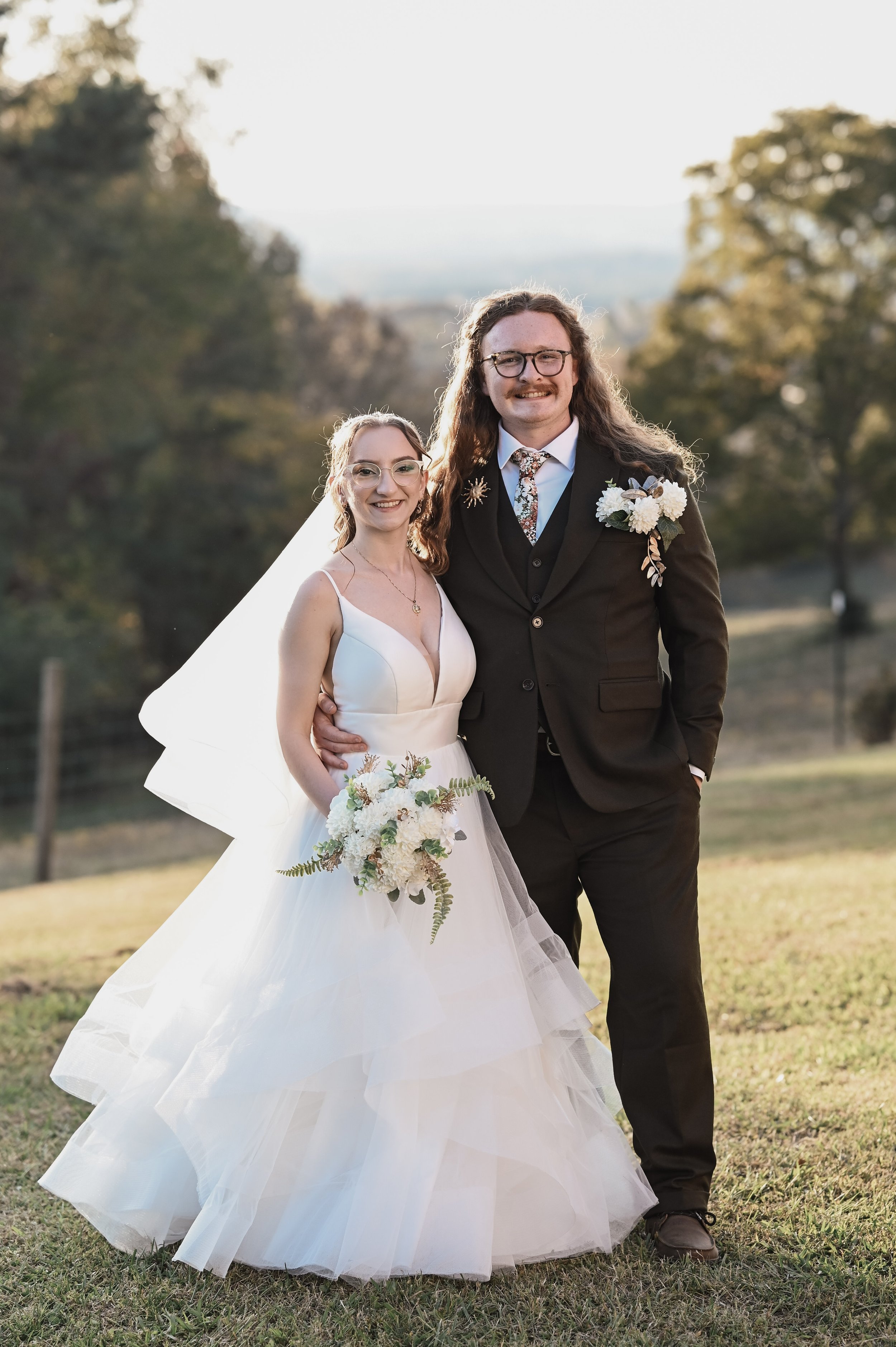 A bride and groom posing outdoors on their wedding day, smiling, with trees and a grassy field in the background during sunset.