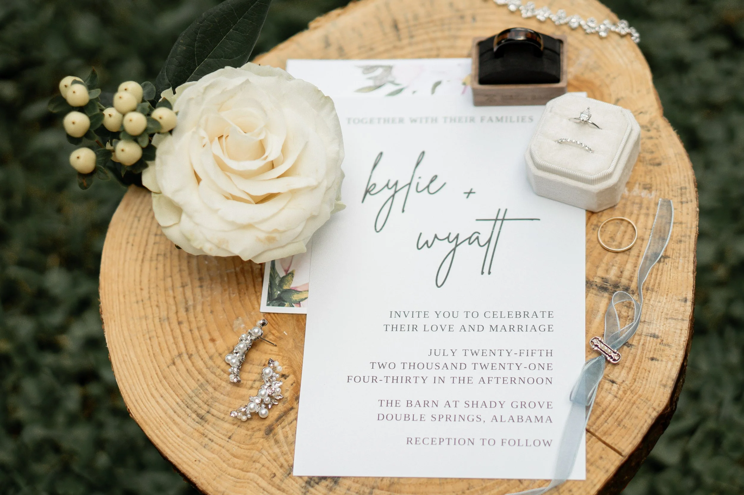 Wedding invitation card surrounded by a white rose, earrings, a wedding band, a ring, and a necklace on a wooden surface.