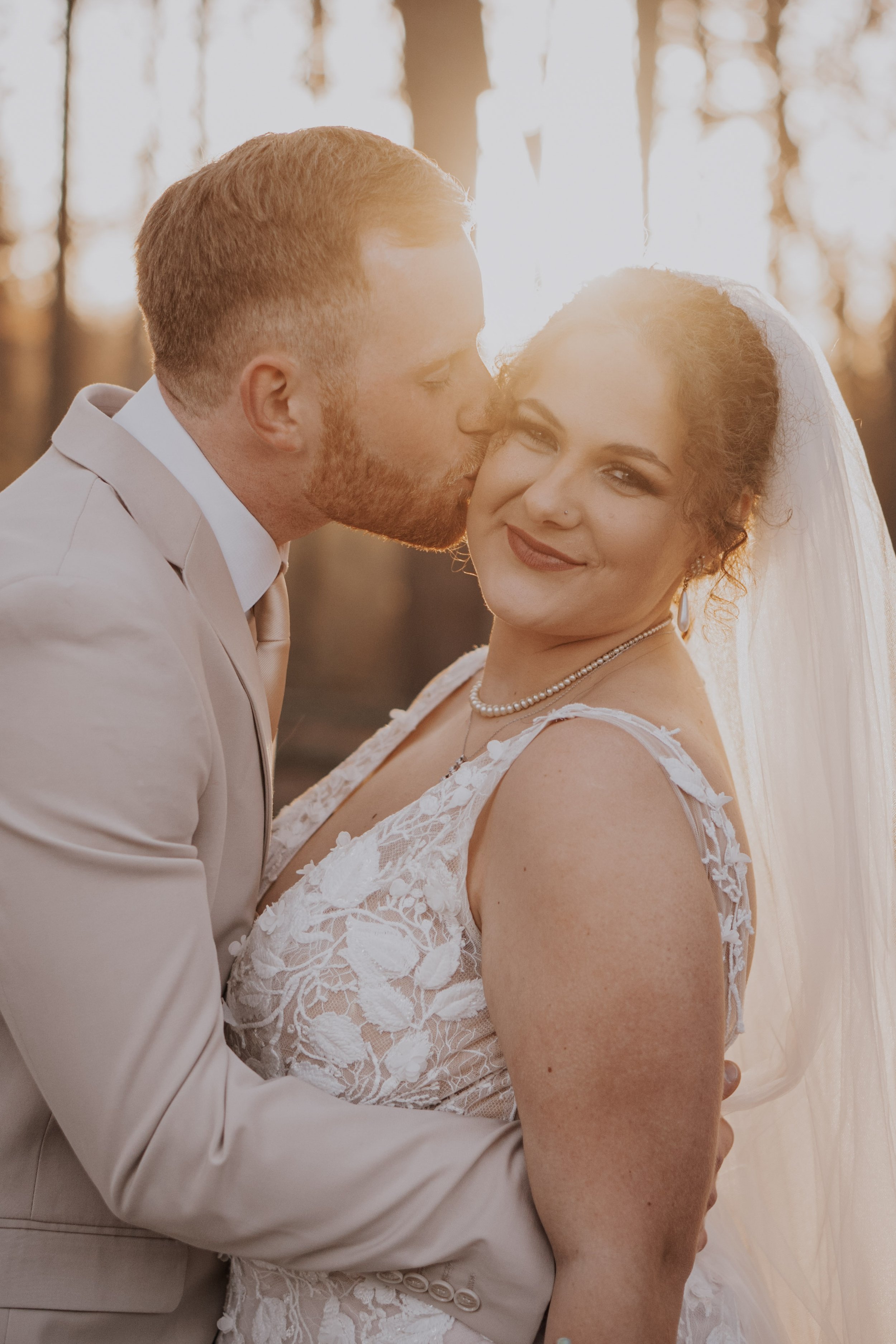 A bride and groom sharing an intimate moment outdoors during sunset, with the groom kissing the bride on the cheek.