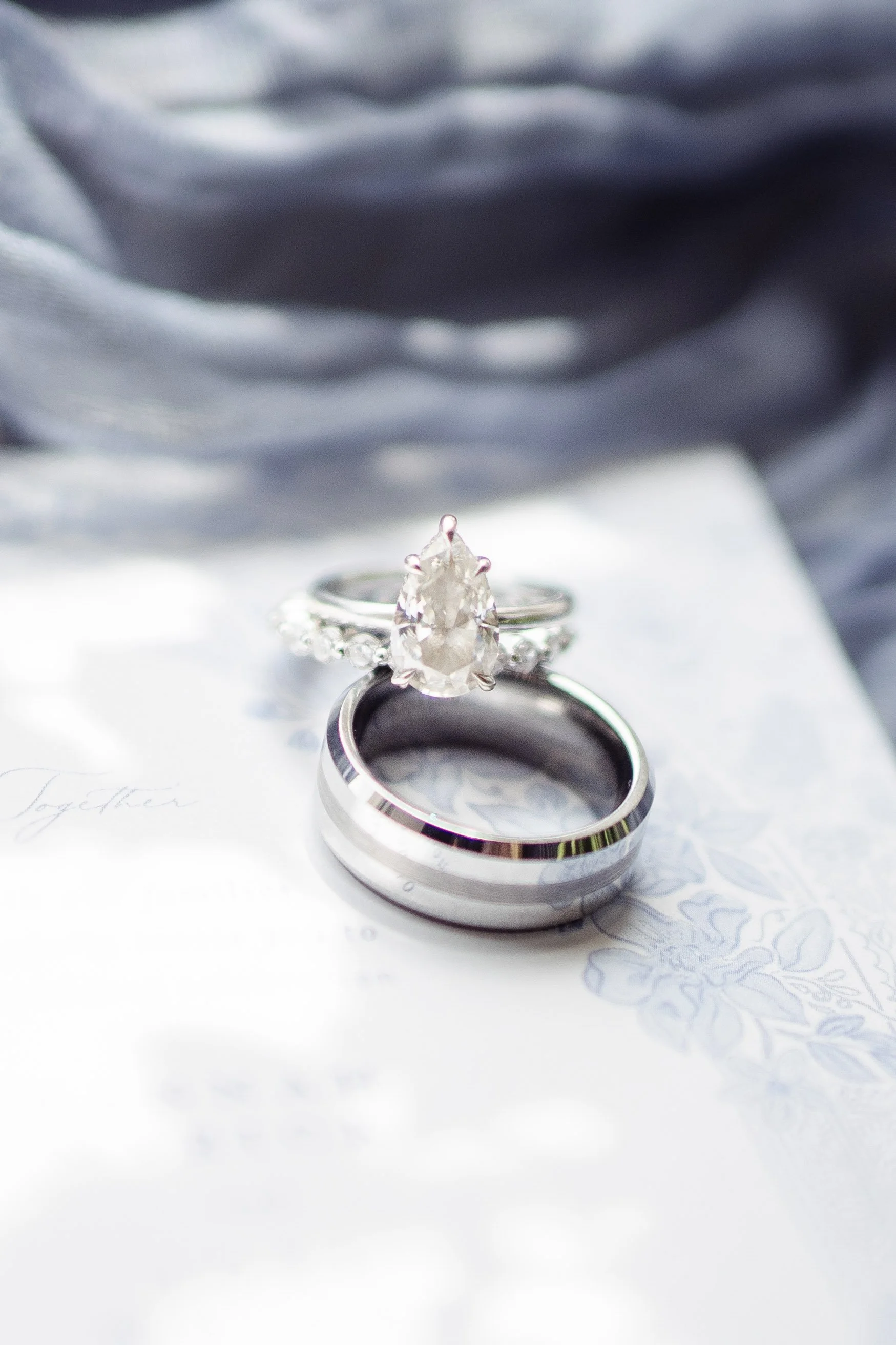 Close-up of two wedding rings, one with a pear-shaped diamond engagement ring and a simple band, resting on a document with floral design.