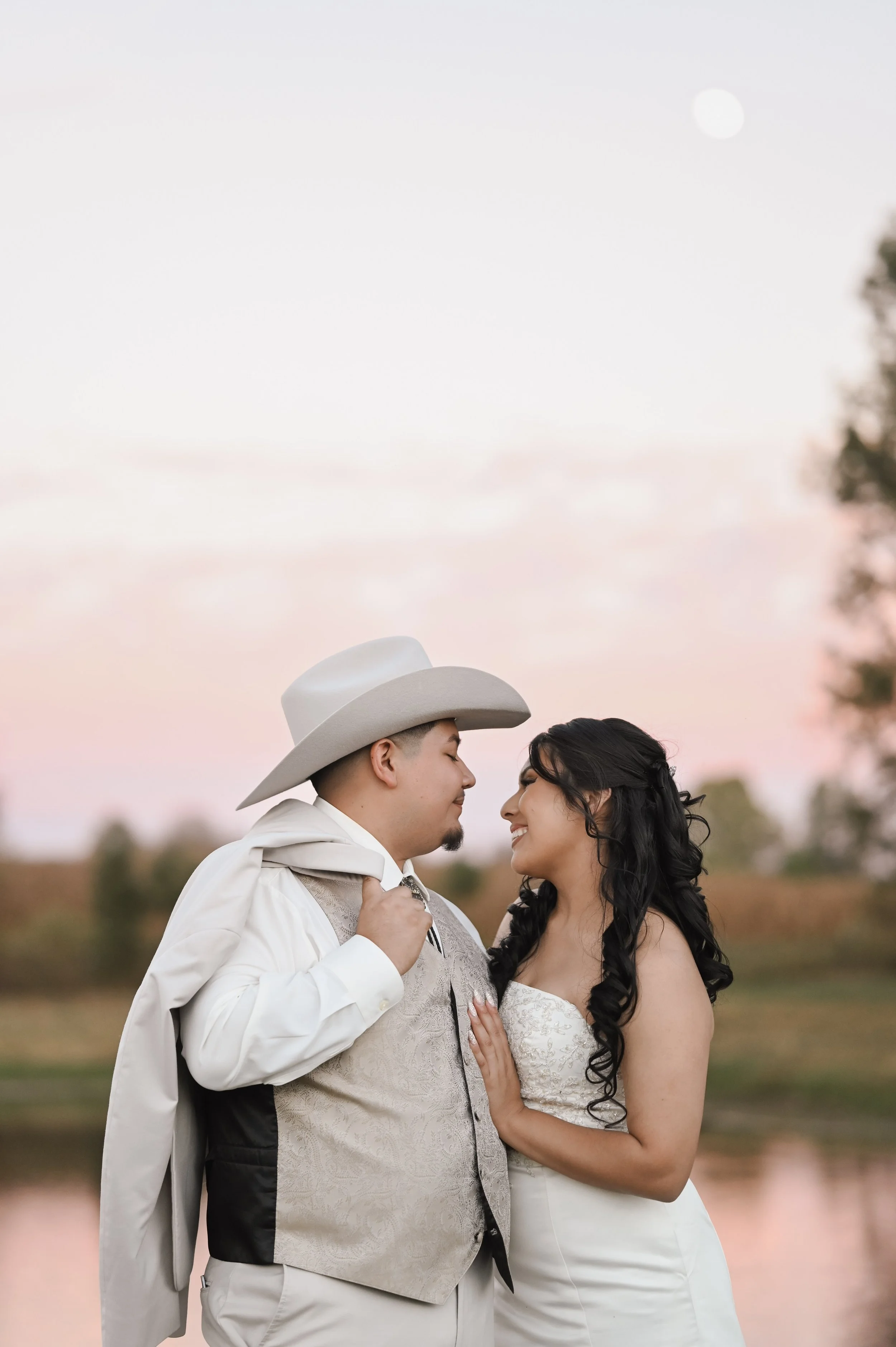 A couple dressed in wedding attire standing close together outdoors during sunset, with a pink sky and a body of water in the background.