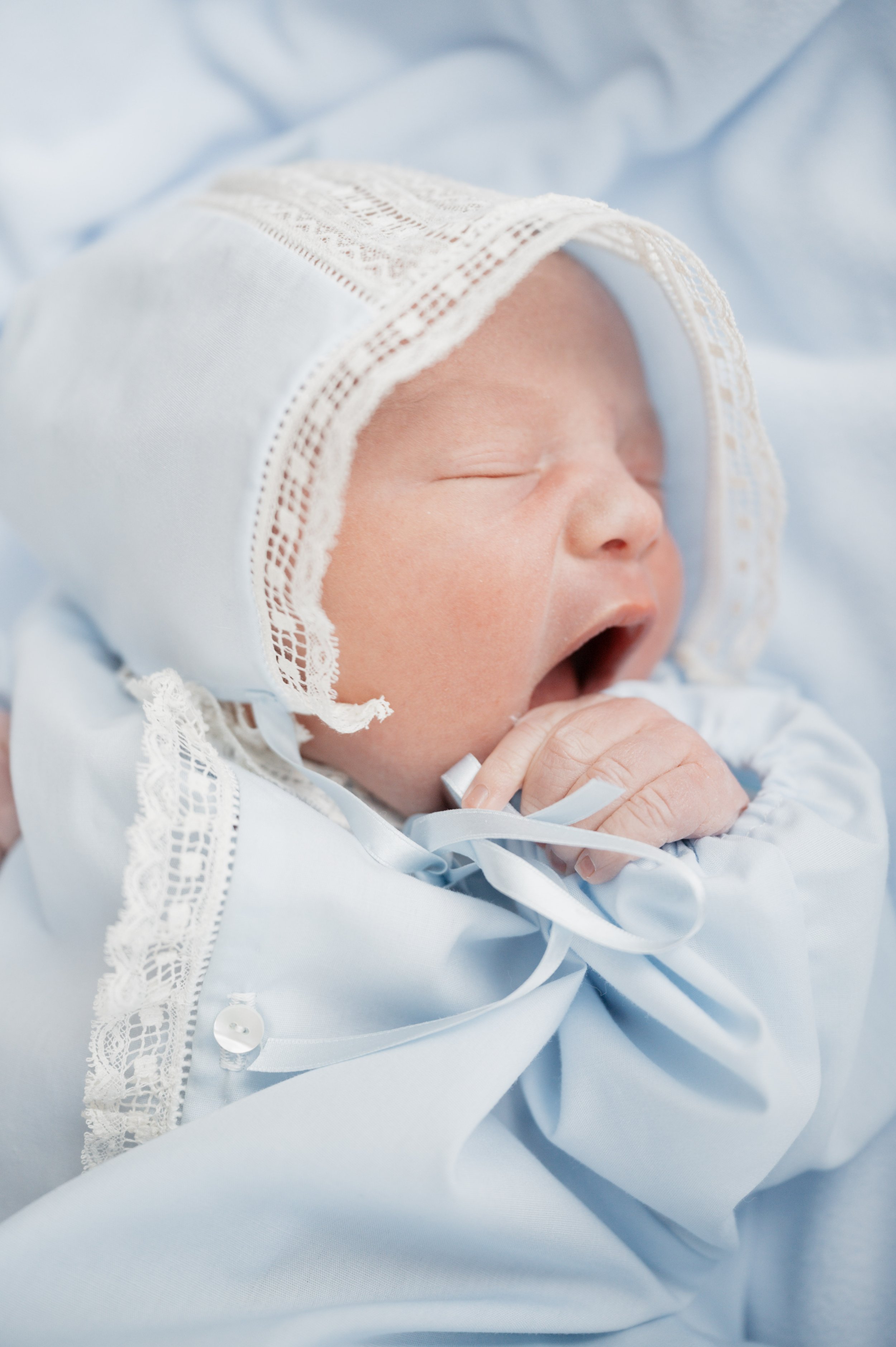 A newborn baby wearing a light blue outfit and a lace-edged bonnet, yawning and sucking on a finger.