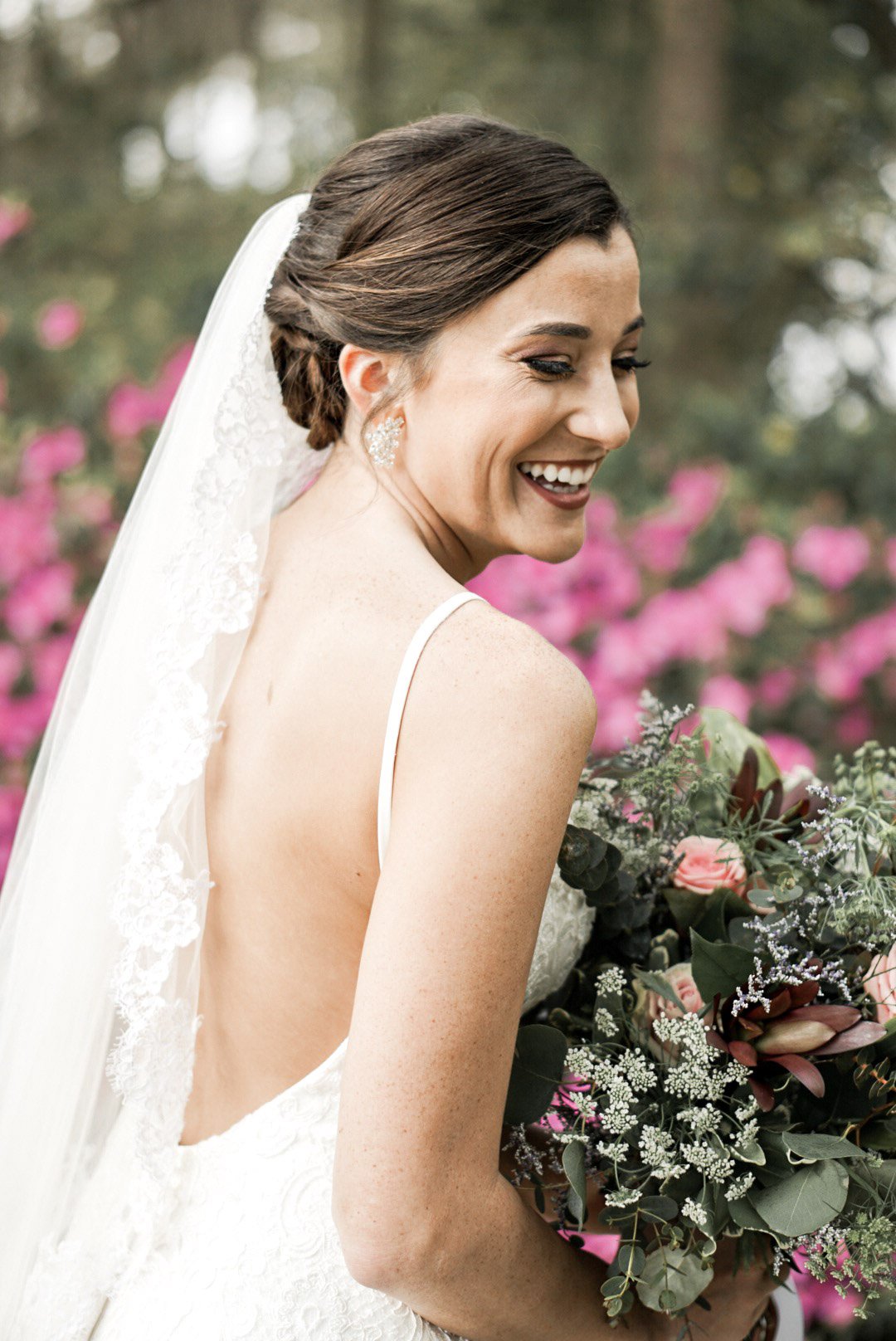 A smiling bride in a white wedding dress holding a bouquet of pink and green flowers, outdoors with pink flowers in the background.