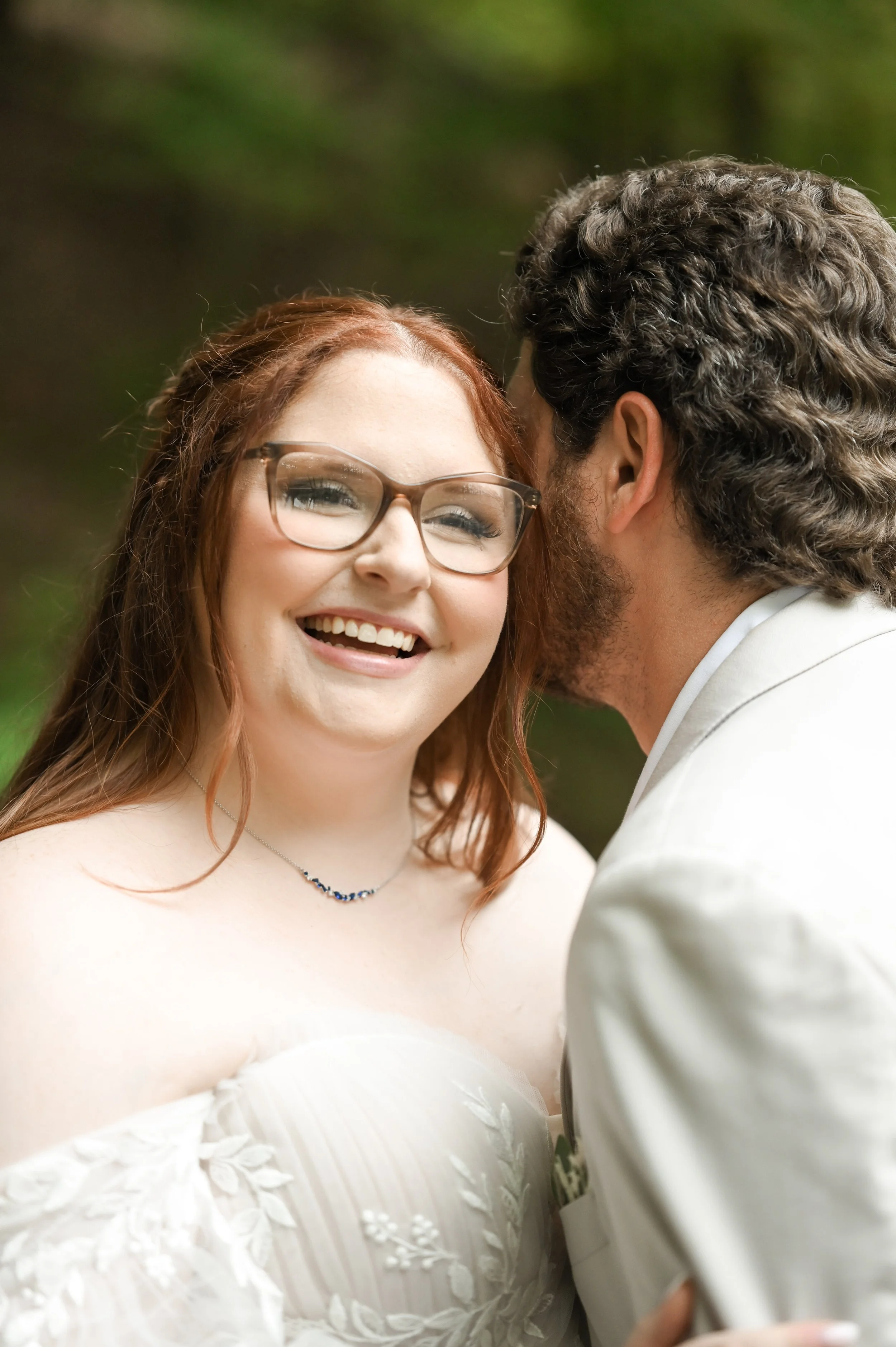 A smiling woman with red hair and glasses in a wedding dress being whispered to by a man in a white suit, outdoors with blurred green foliage in the background.