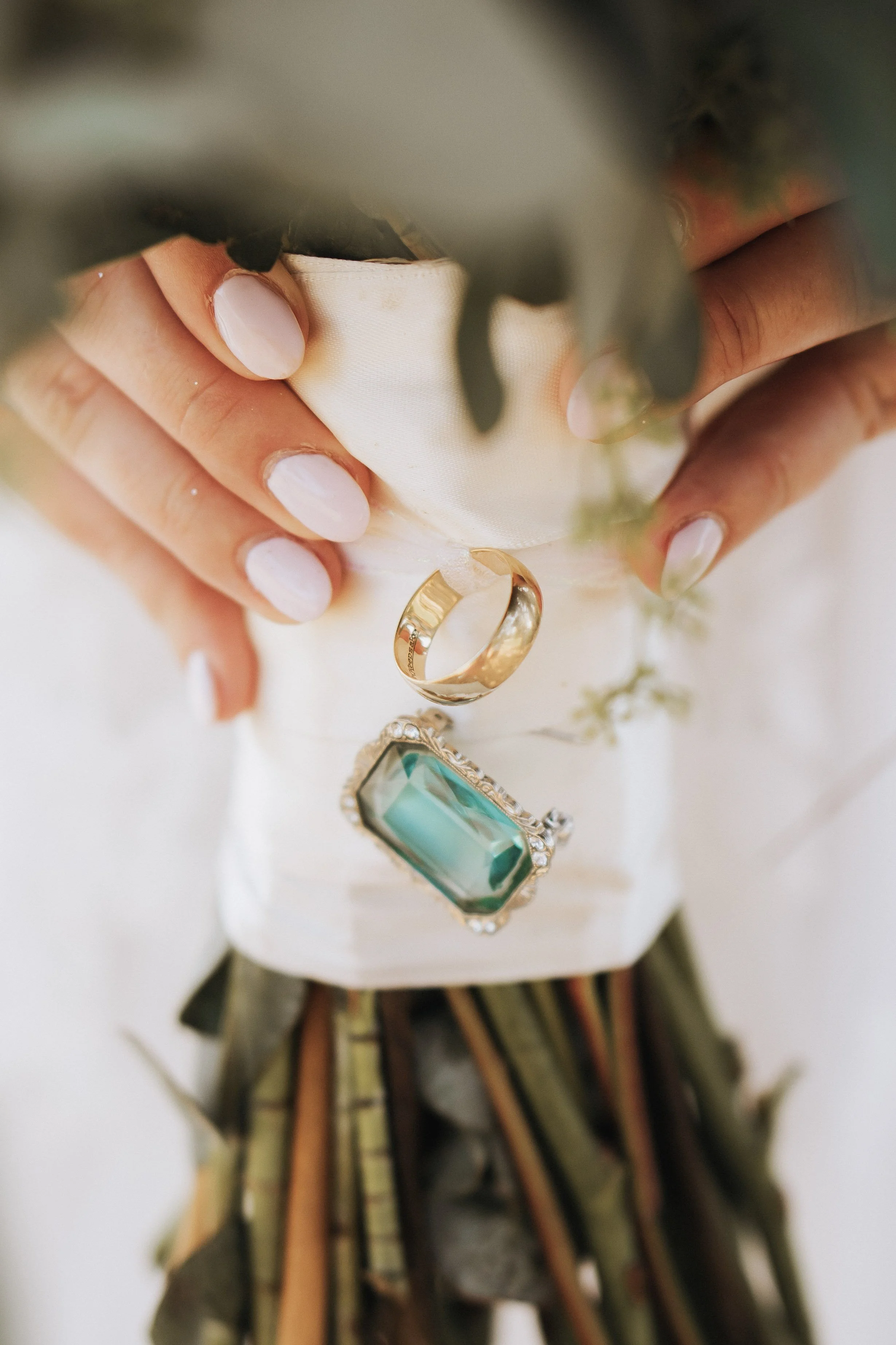 Close-up of a person's hand holding a small bouquet with a gold ring and a large gemstone ring resting on a white cloth.