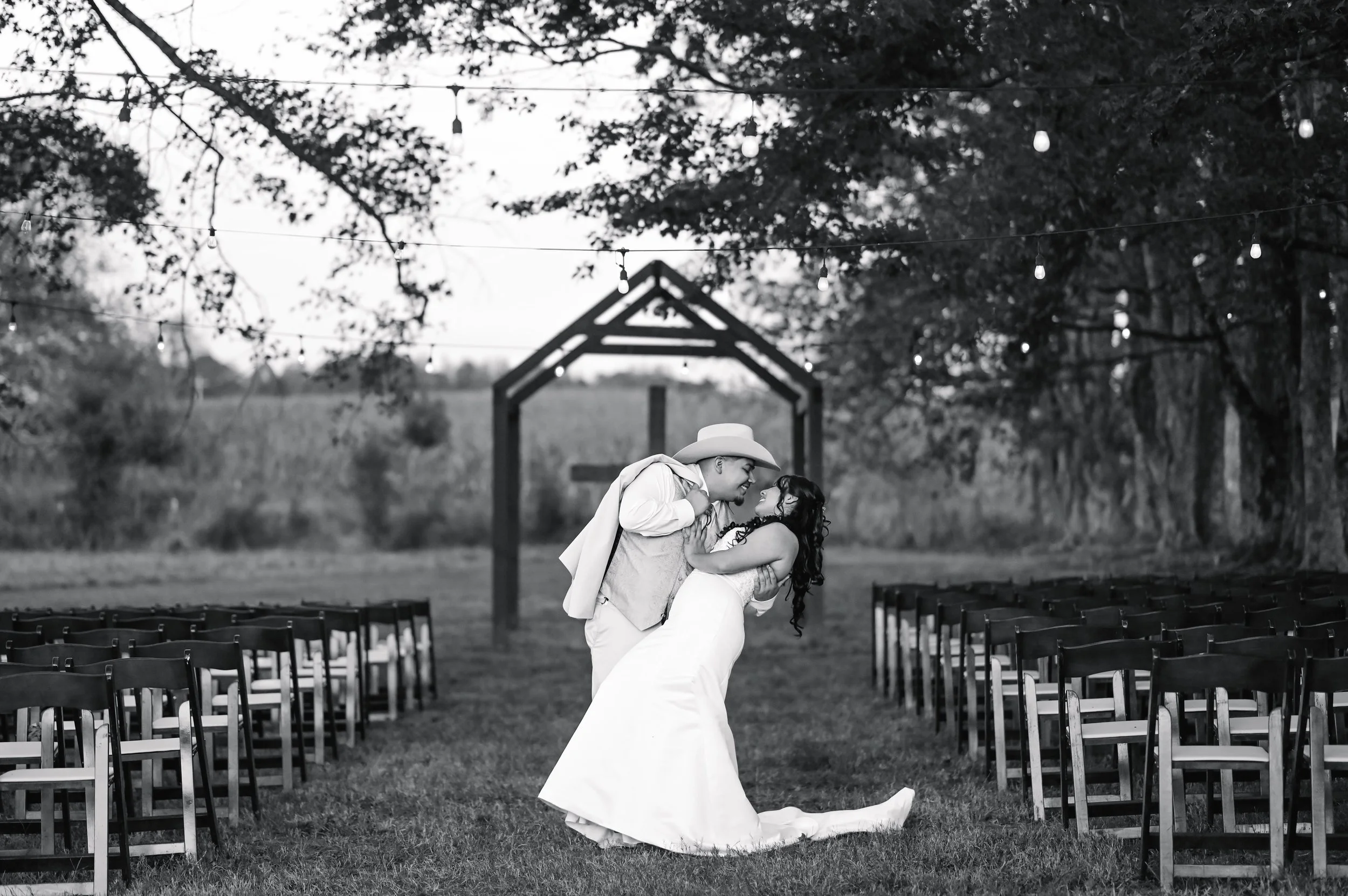Couple dancing at an outdoor wedding ceremony, surrounded by chairs and decorative lights, with trees and a wooden arch in the background.