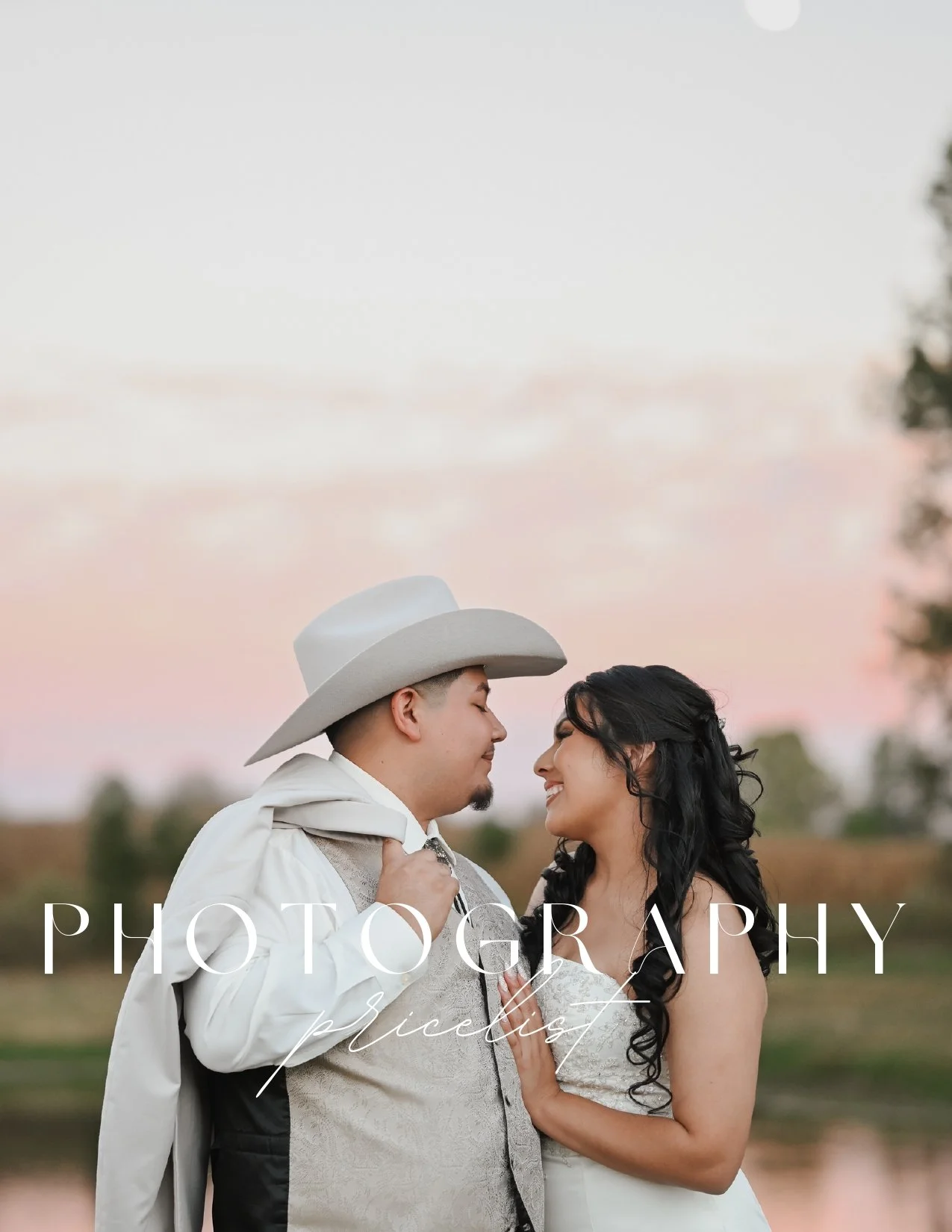 Couple dressed in formal attire, smiling and close together outdoors at sunset or sunrise with a lake and trees in the background.