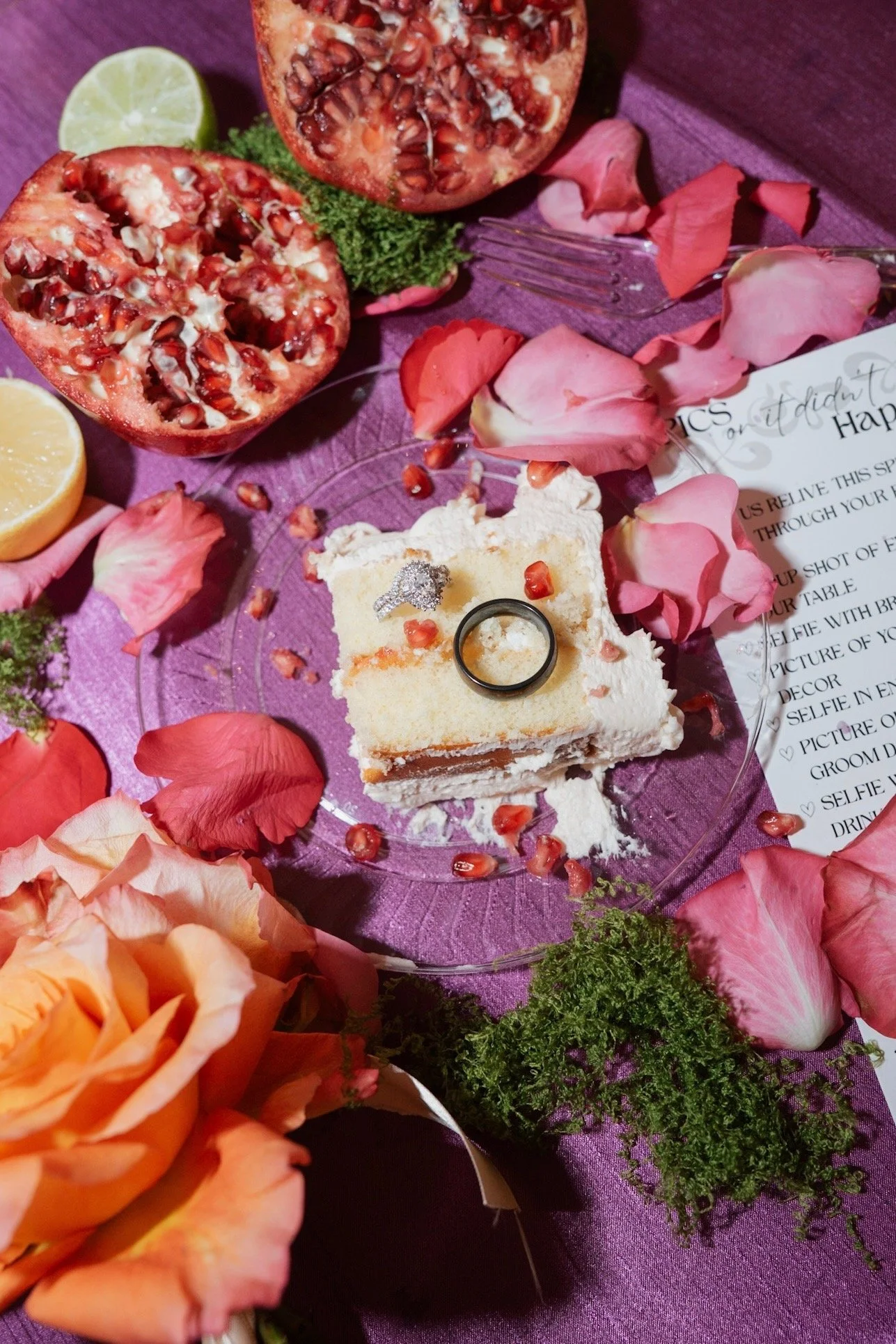 A dessert table with pomegranate halves, lemon wedges, pink rose petals, and a layered cake with a black ring on top, all arranged on a purple surface with decorative greenery and a partially visible printed message.