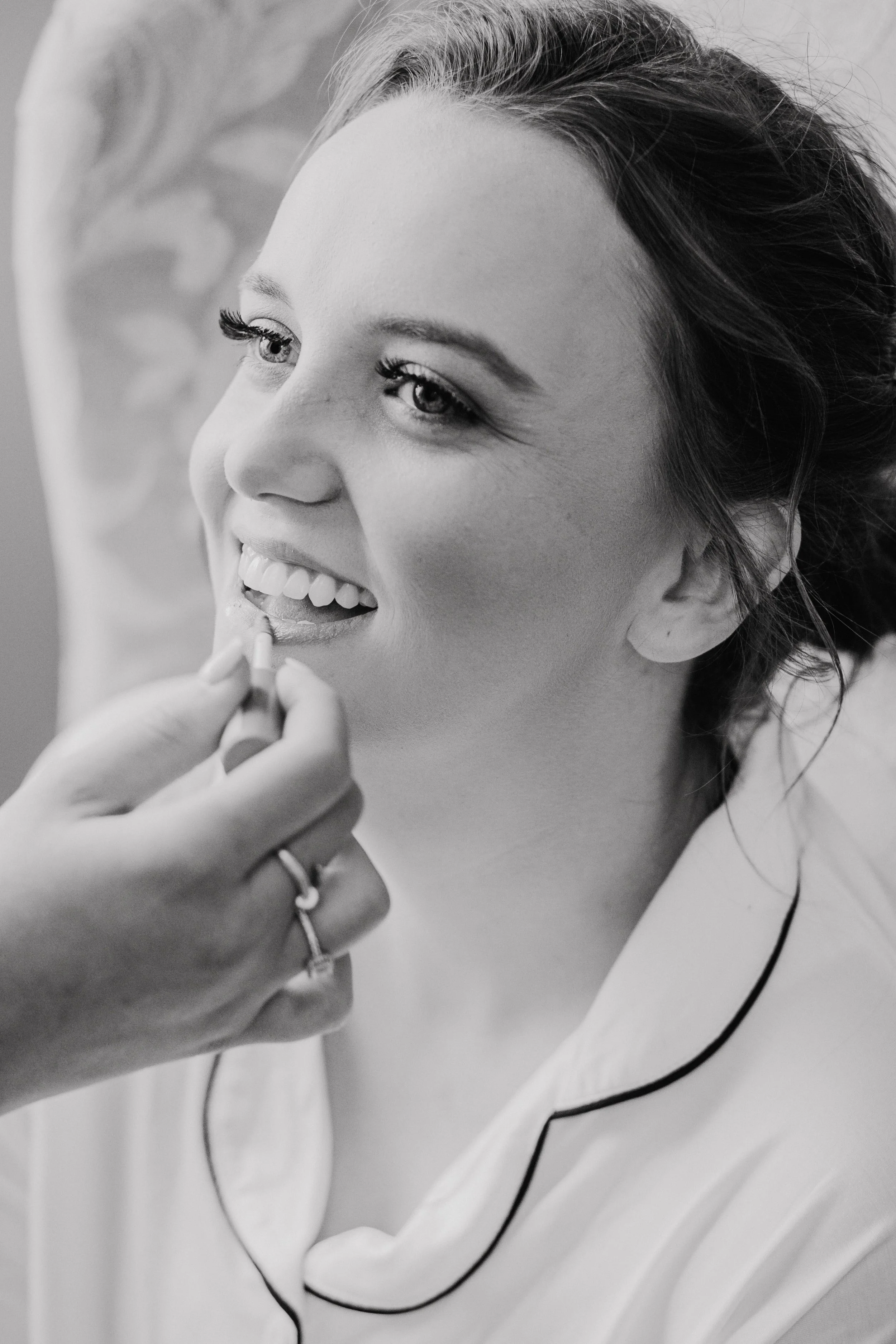 A woman having lipstick applied to her lips by another person, smiling with visible teeth, wearing a satin robe with black piping, in a close-up black and white photo.