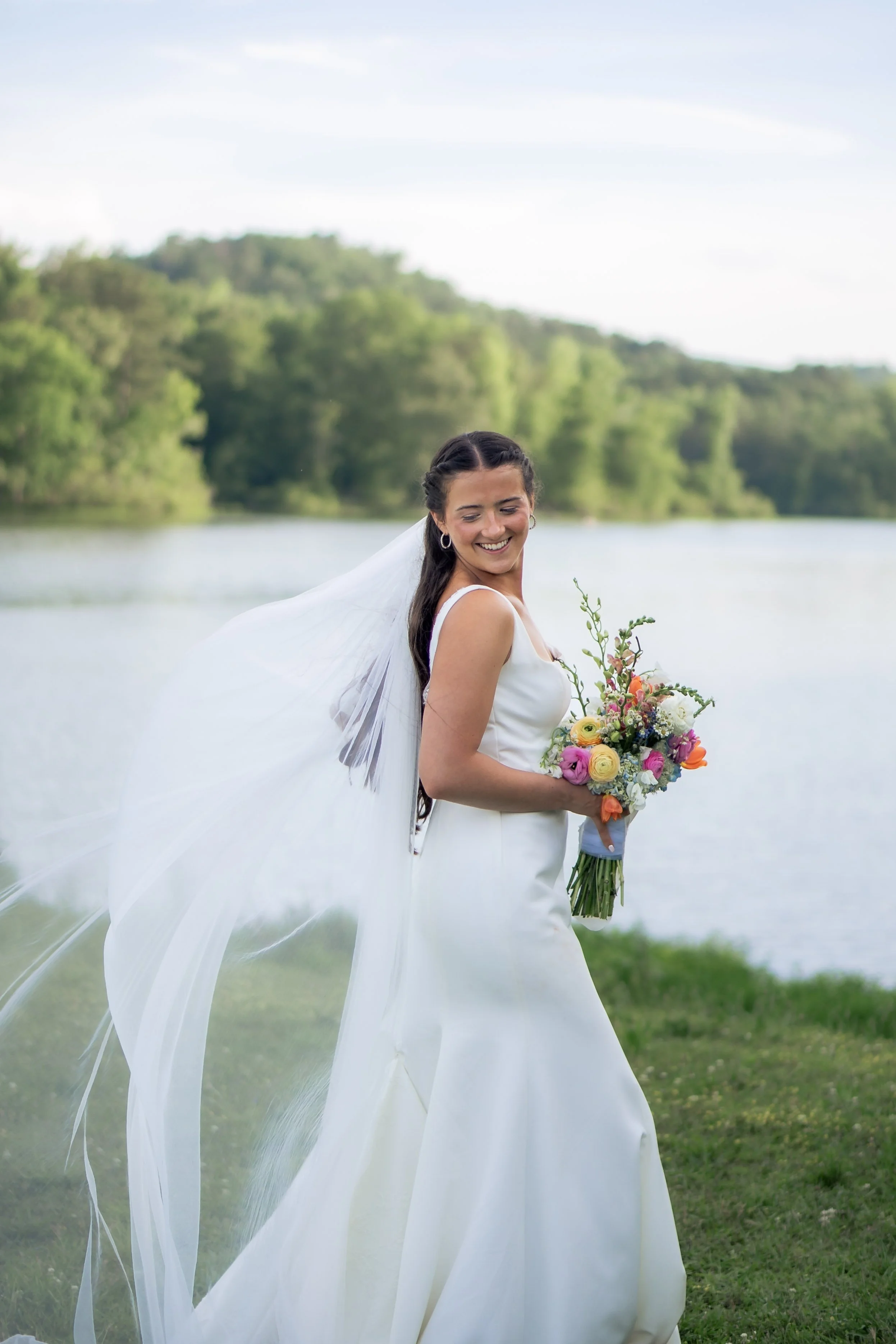 A bride in a white wedding dress holding a colorful bouquet, standing near a river with green trees and a blue sky in the background.
