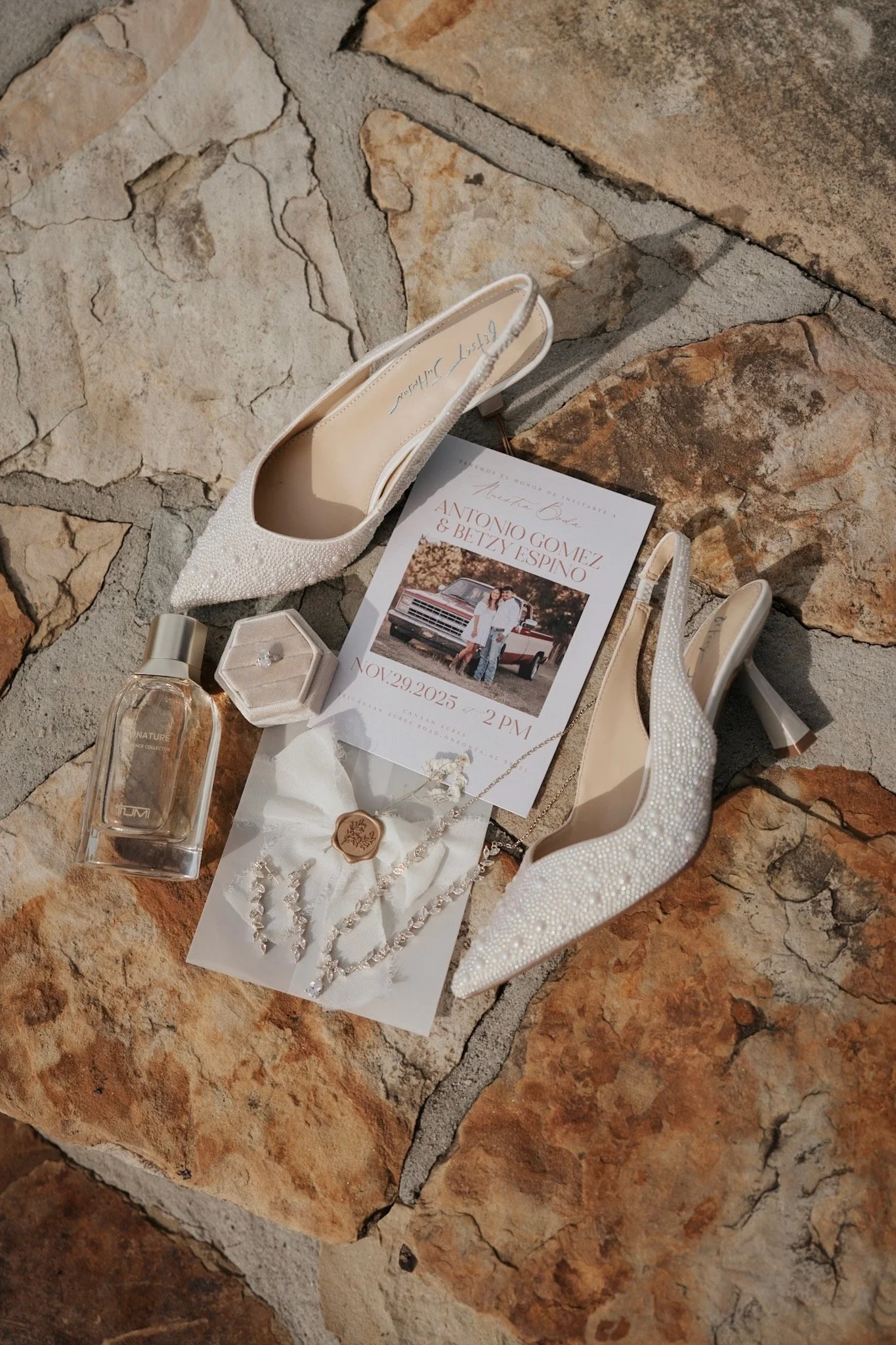 A pair of white, pointed-toe high heels with pearl embellishments, a small perfume bottle, a jewelry box, a wedding invitation, a wax seal stamp, a necklace, and a bracelet are arranged on a rustic stone floor.