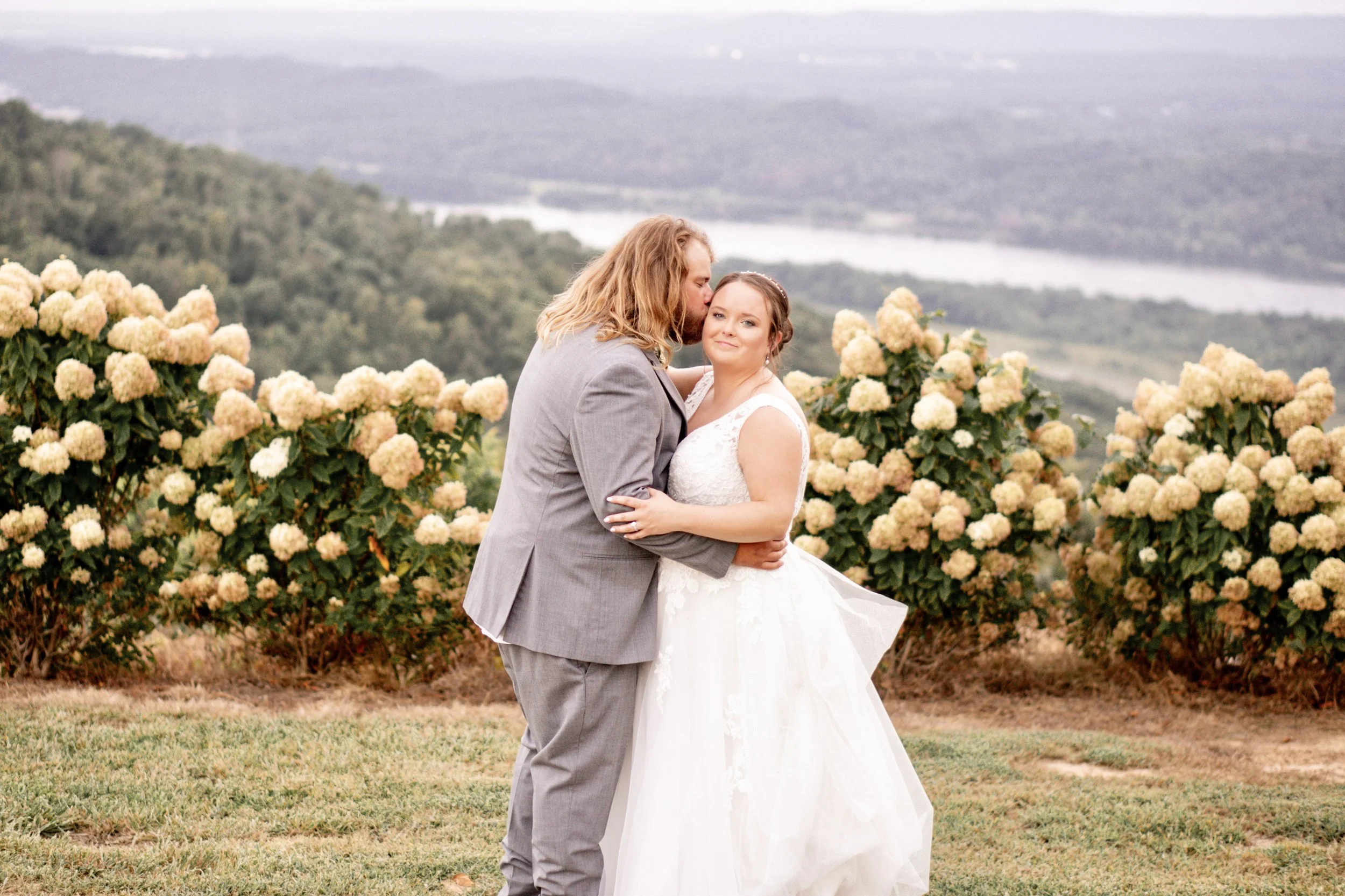 A couple, dressed in wedding attire, sharing an intimate moment outdoors among blooming hydrangea bushes with a scenic landscape and river in the background.