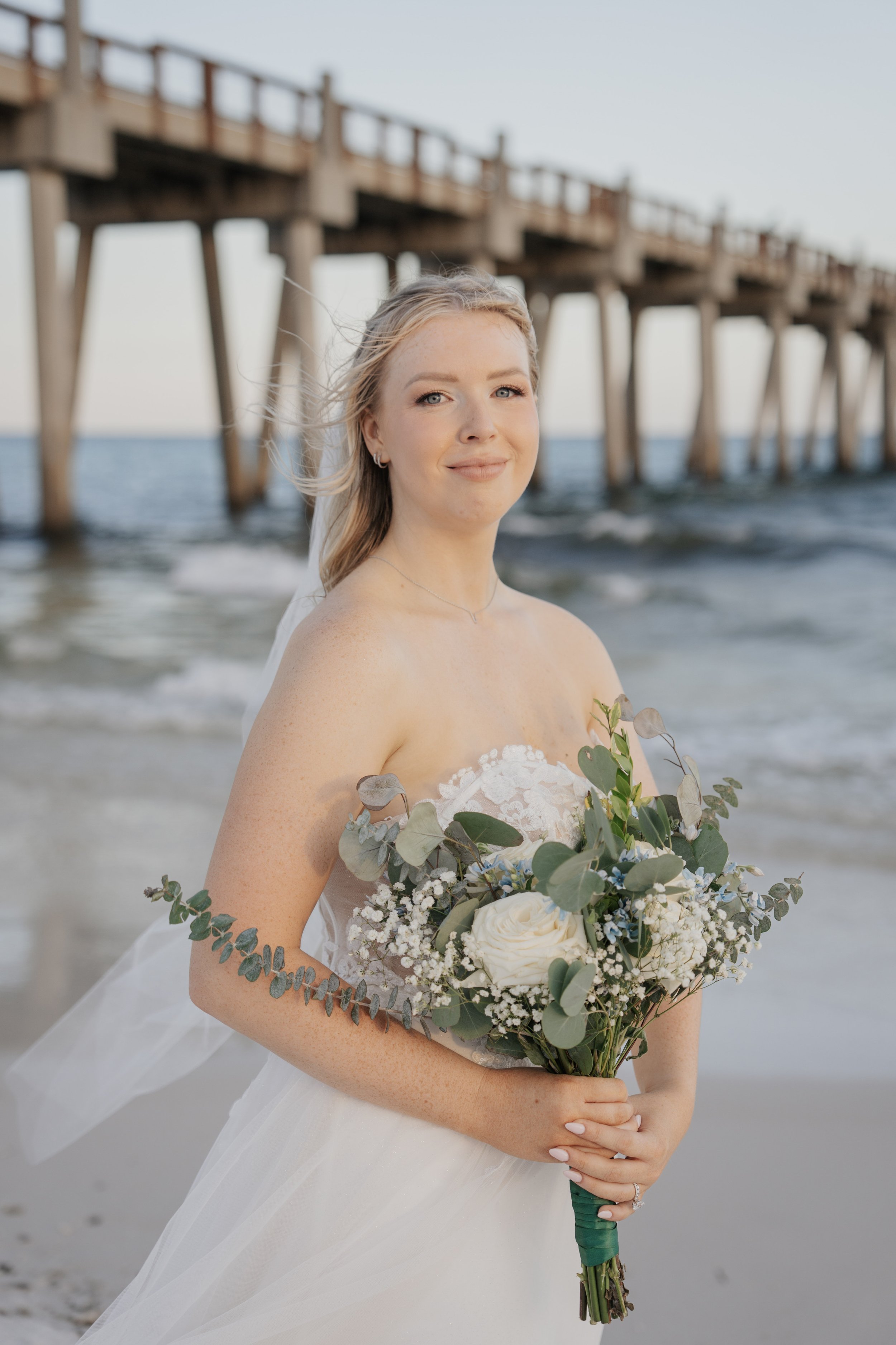 A bride standing on the beach holding a bouquet of white flowers and greenery, with a wooden pier in the background.