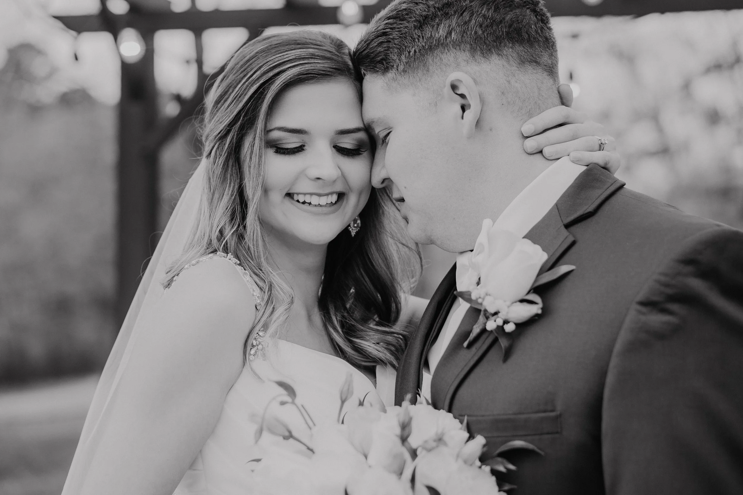 A bride and groom share a intimate moment on their wedding day, with their foreheads touching and eyes closed, smiling gently. The bride is wearing a wedding dress with lace details and a veil, while the groom is dressed in a suit with a boutonniere.