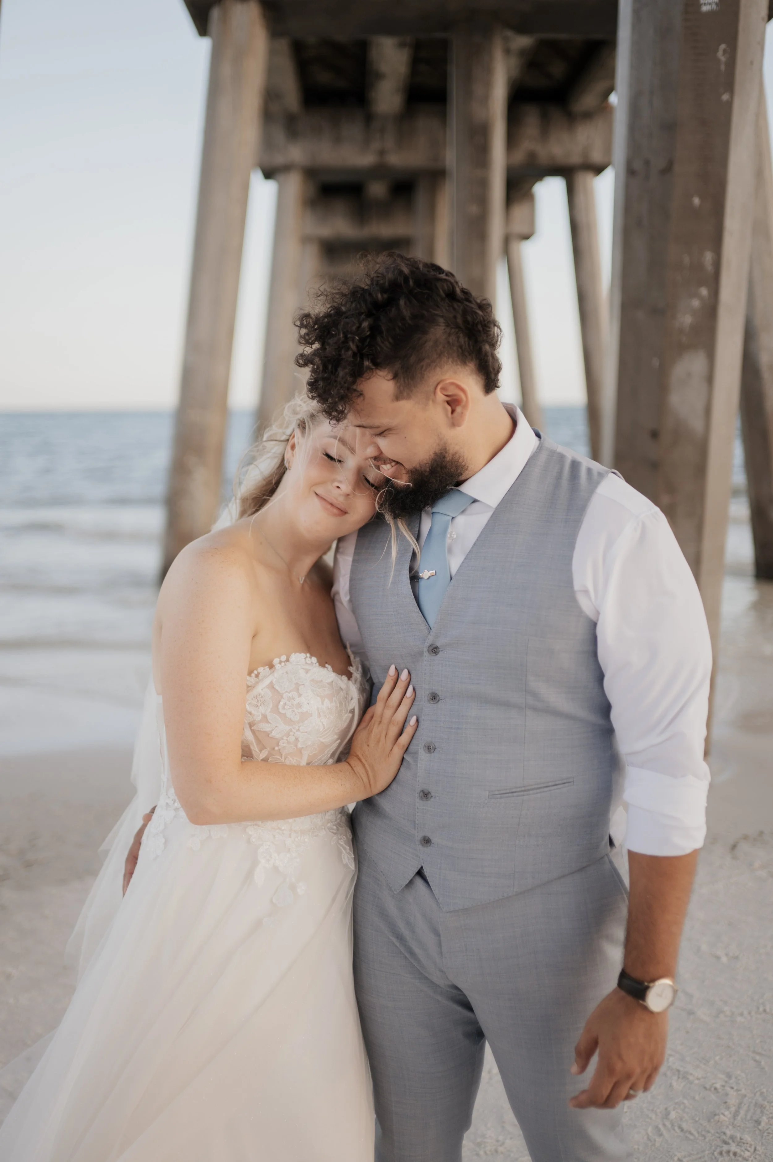 A newlywed couple sharing a tender moment on the beach near a wooden pier, with the bride in a white lace wedding gown and the groom in a light gray suit, both smiling with their foreheads touching.