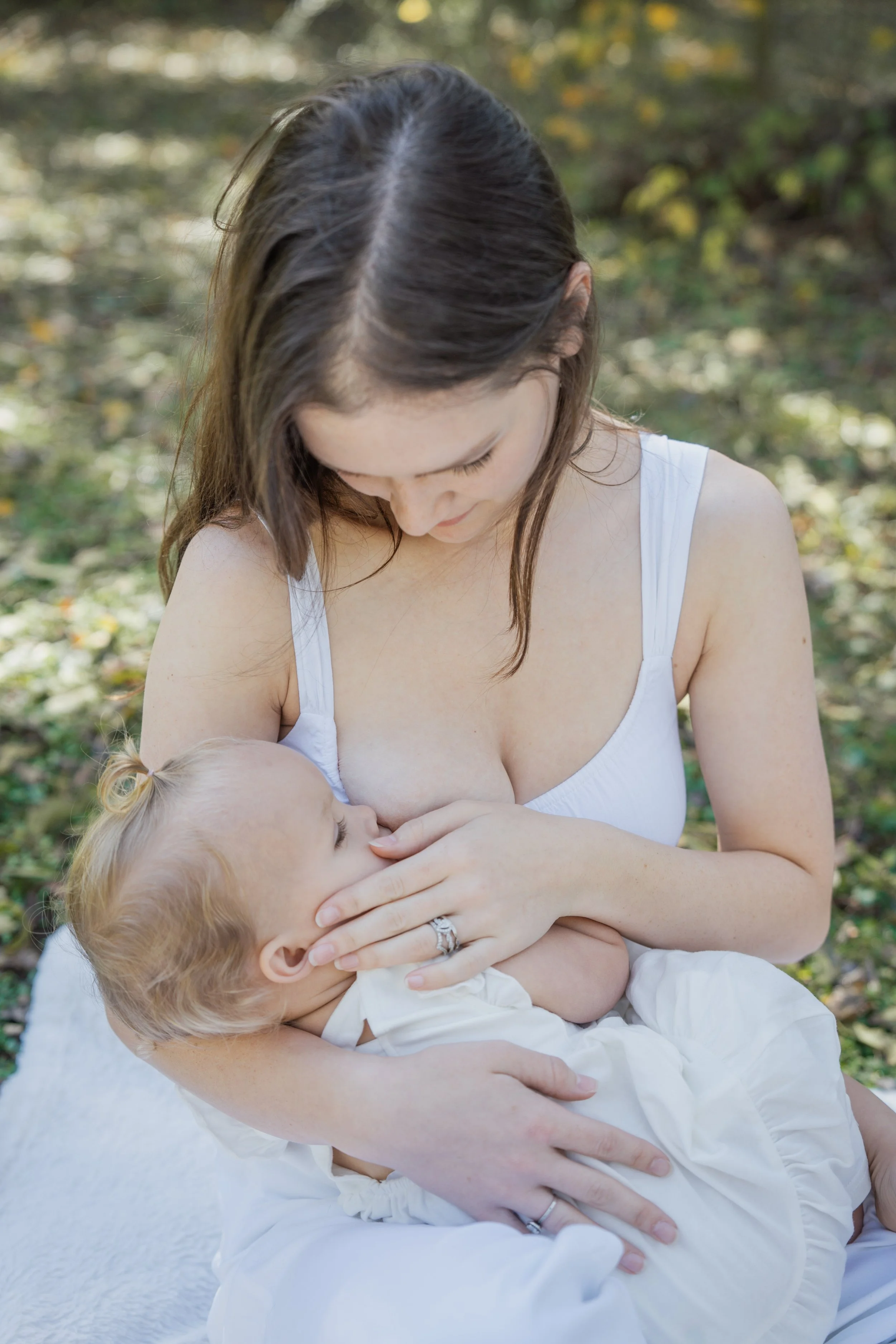 A woman wearing a white tank top breastfeeds a baby outdoors, with a blurred background of trees and leaves.