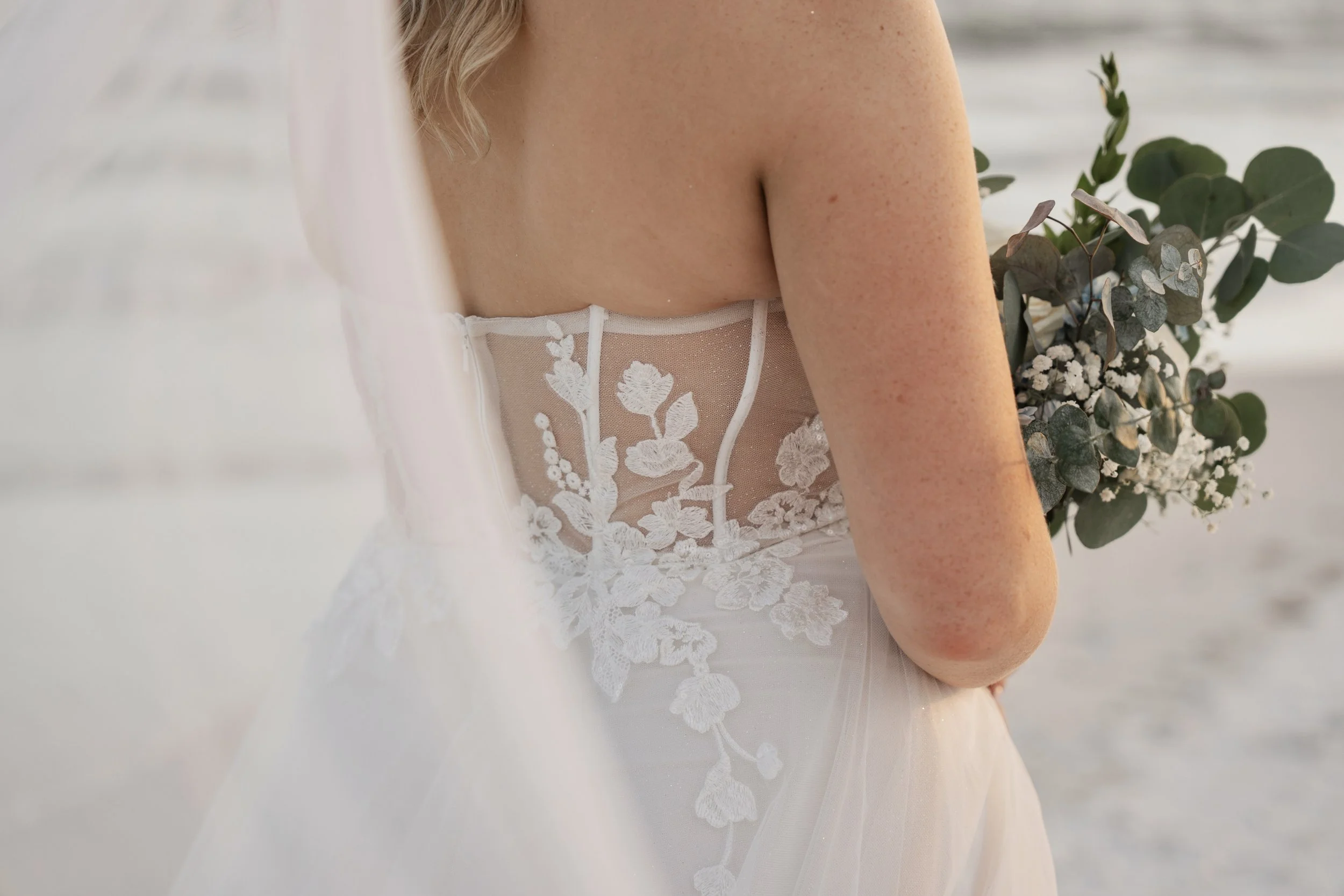 Close-up of a bride in a wedding dress holding a bouquet of white flowers and greenery, with a focus on the intricate lace detailing on the back of her dress.