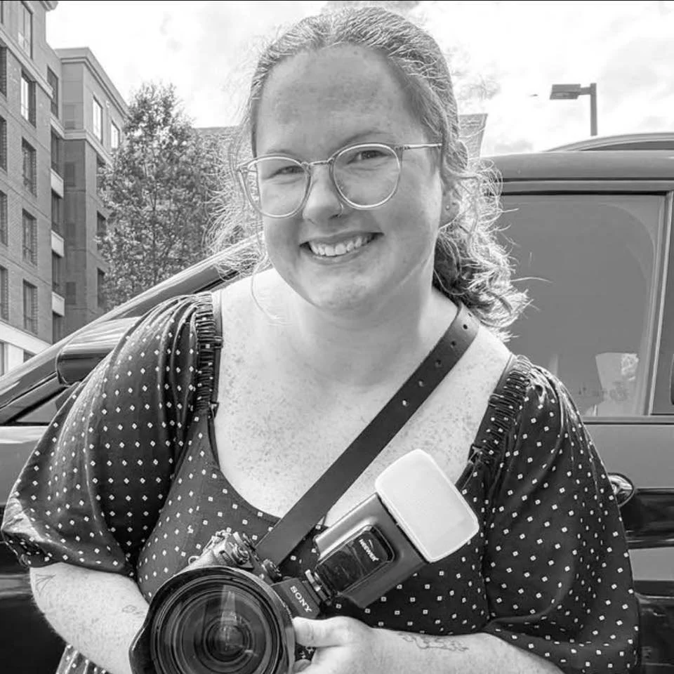 Smiling woman with glasses holding camera standing outside near a parked car.