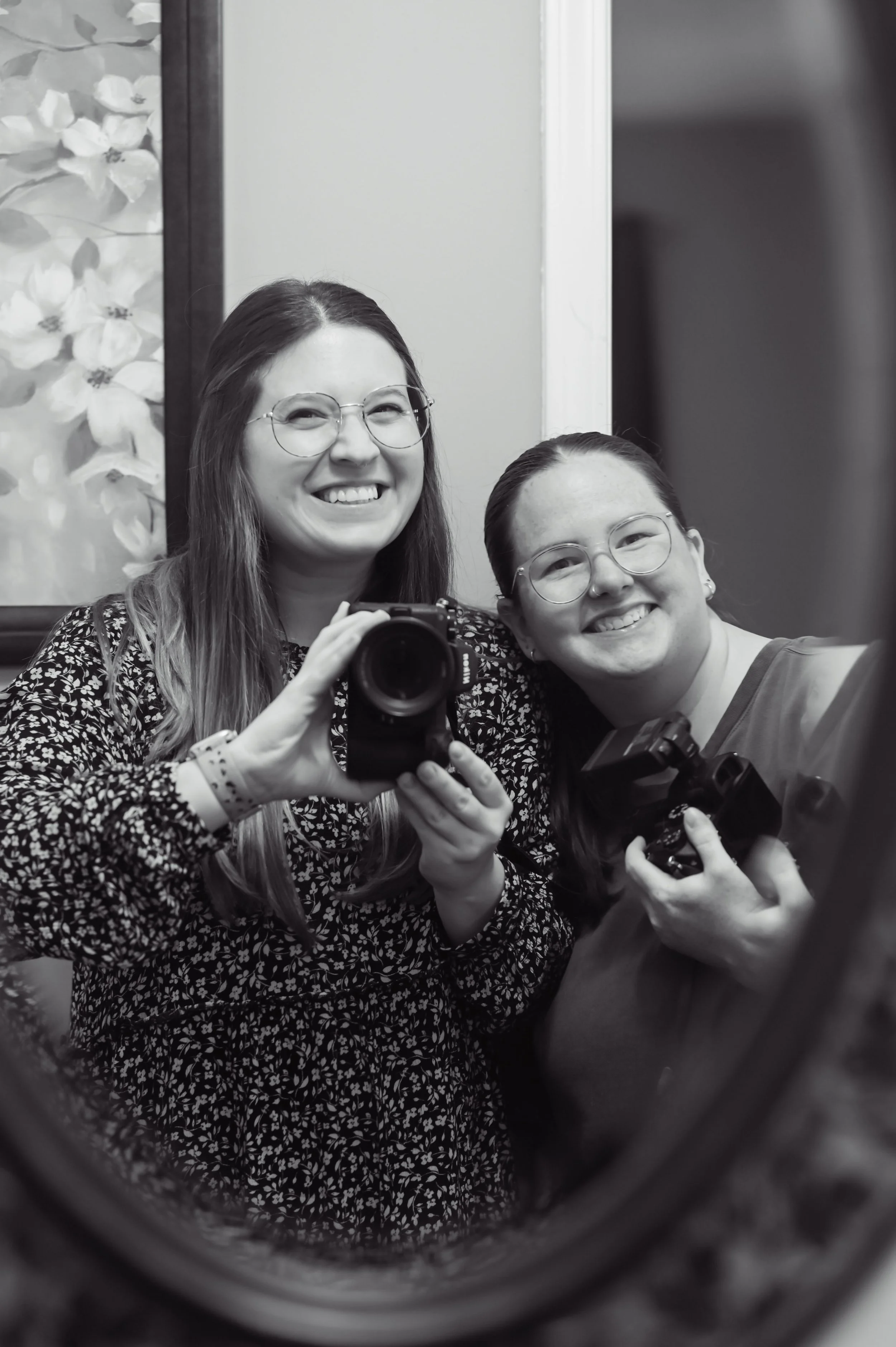 Two women with glasses smiling, taking a selfie in a mirror, holding cameras, in black and white.