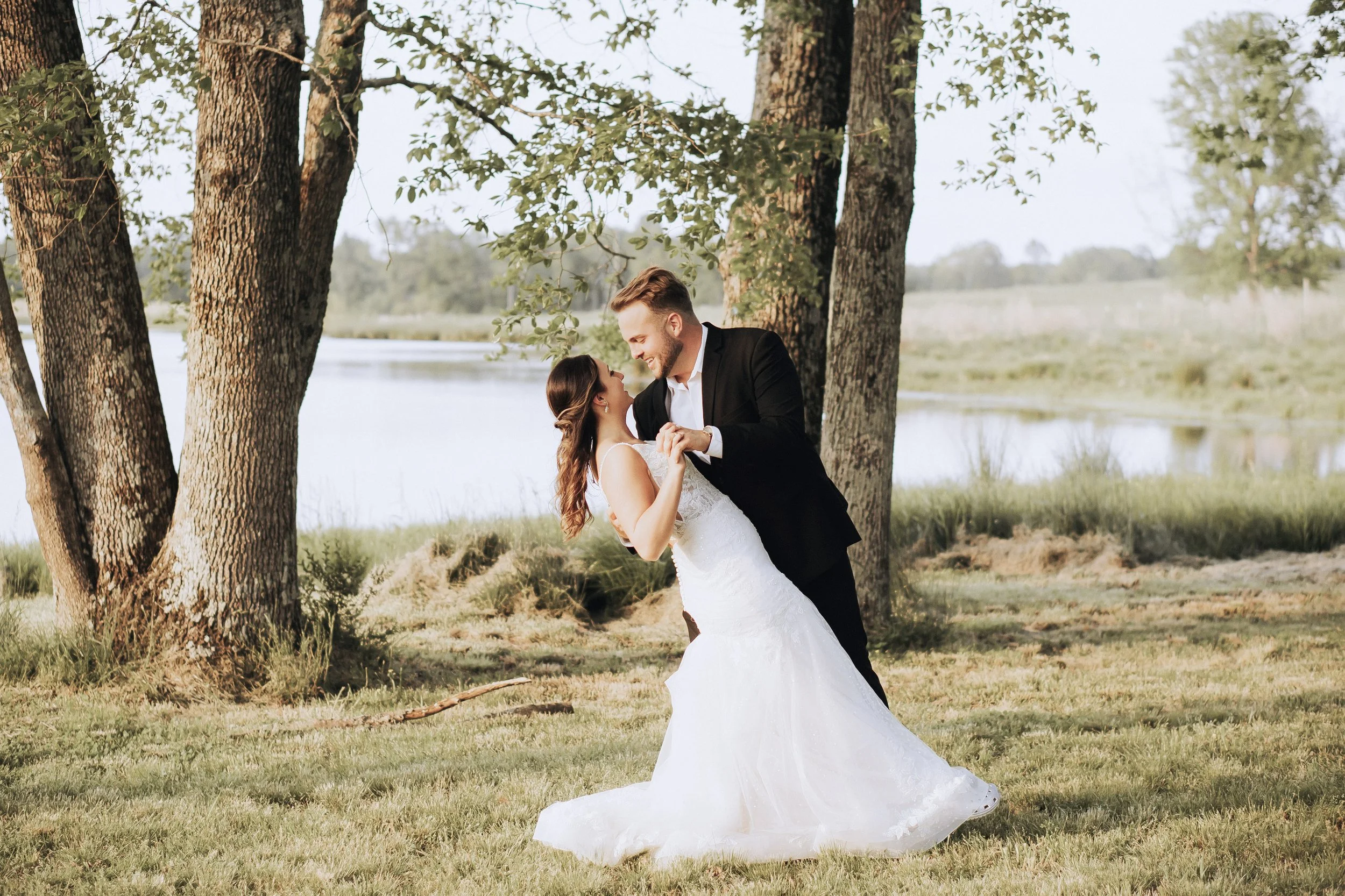 A couple in wedding attire dancing near a lake, surrounded by trees under a clear sky.