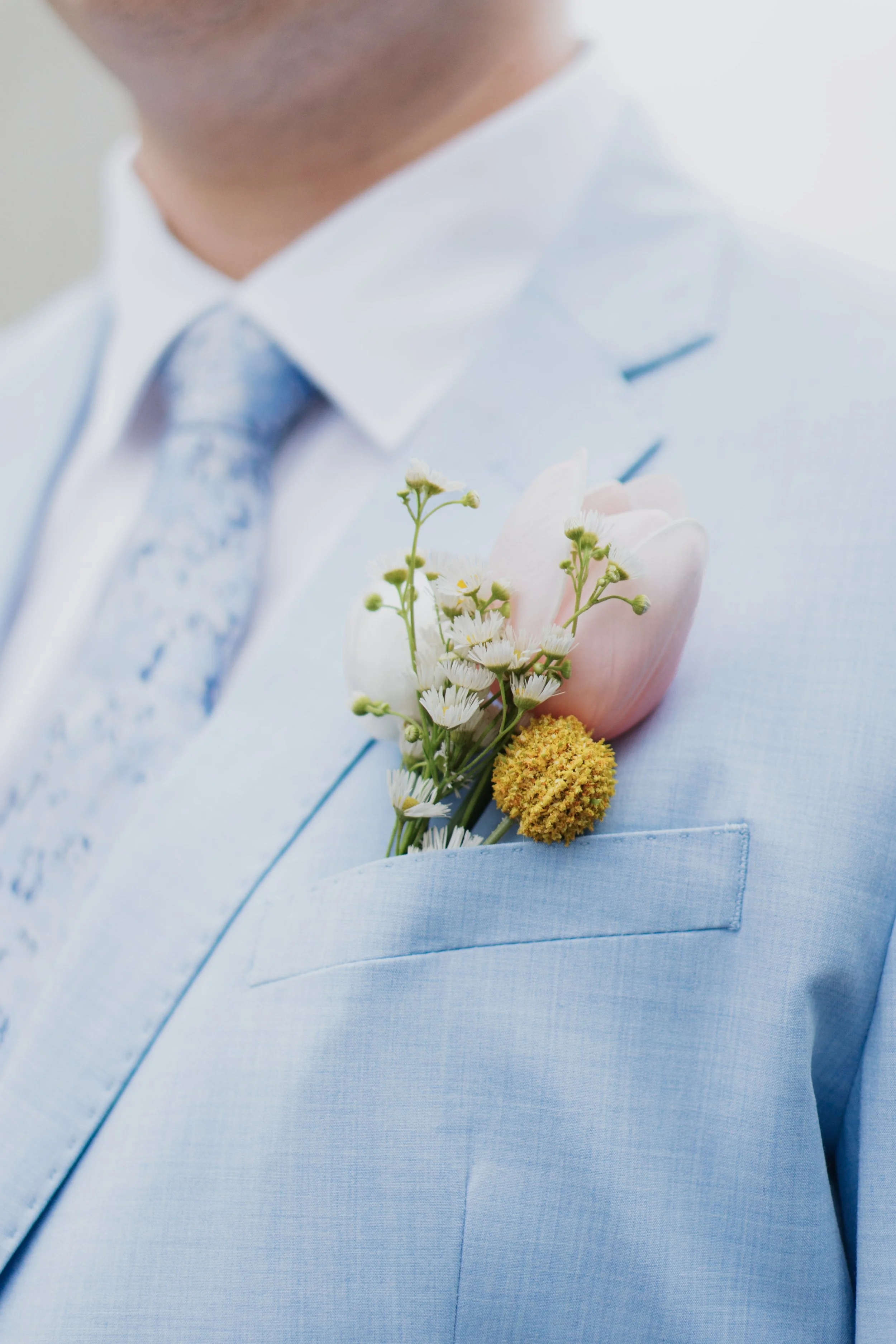 A man in a light blue suit with a light blue patterned tie has a boutonniere with white, pink, and yellow flowers pinned to his chest.