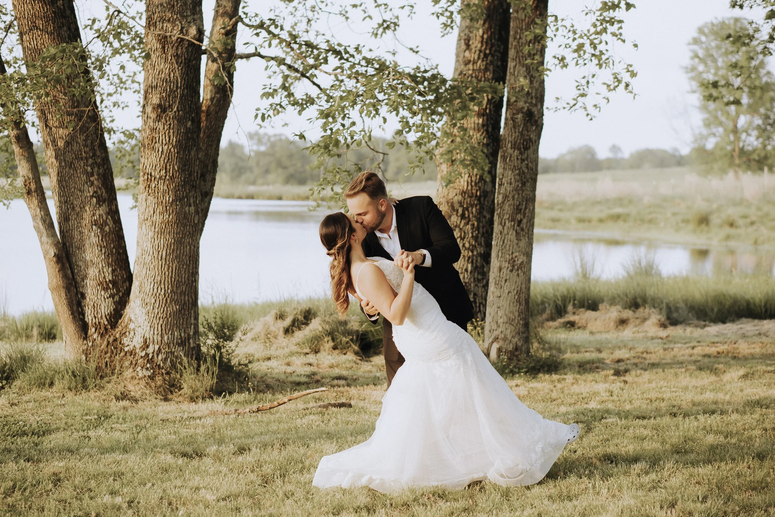 A couple in wedding attire dancing outdoors by a lake, surrounded by trees and grass.