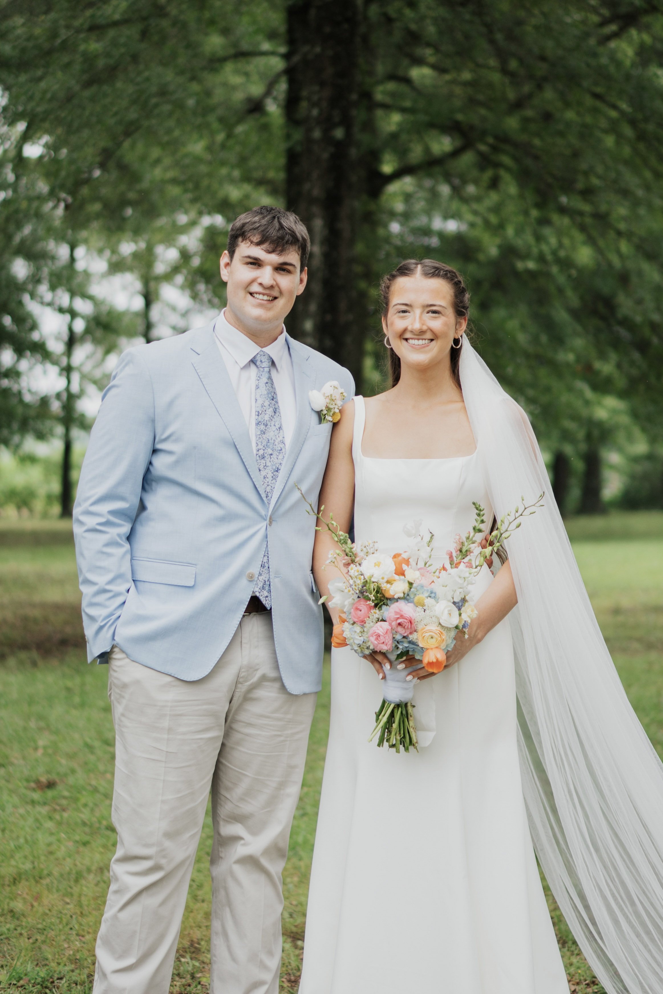 A bride and groom smiling outdoors, with the bride holding a colorful bouquet of flowers, green trees in the background.