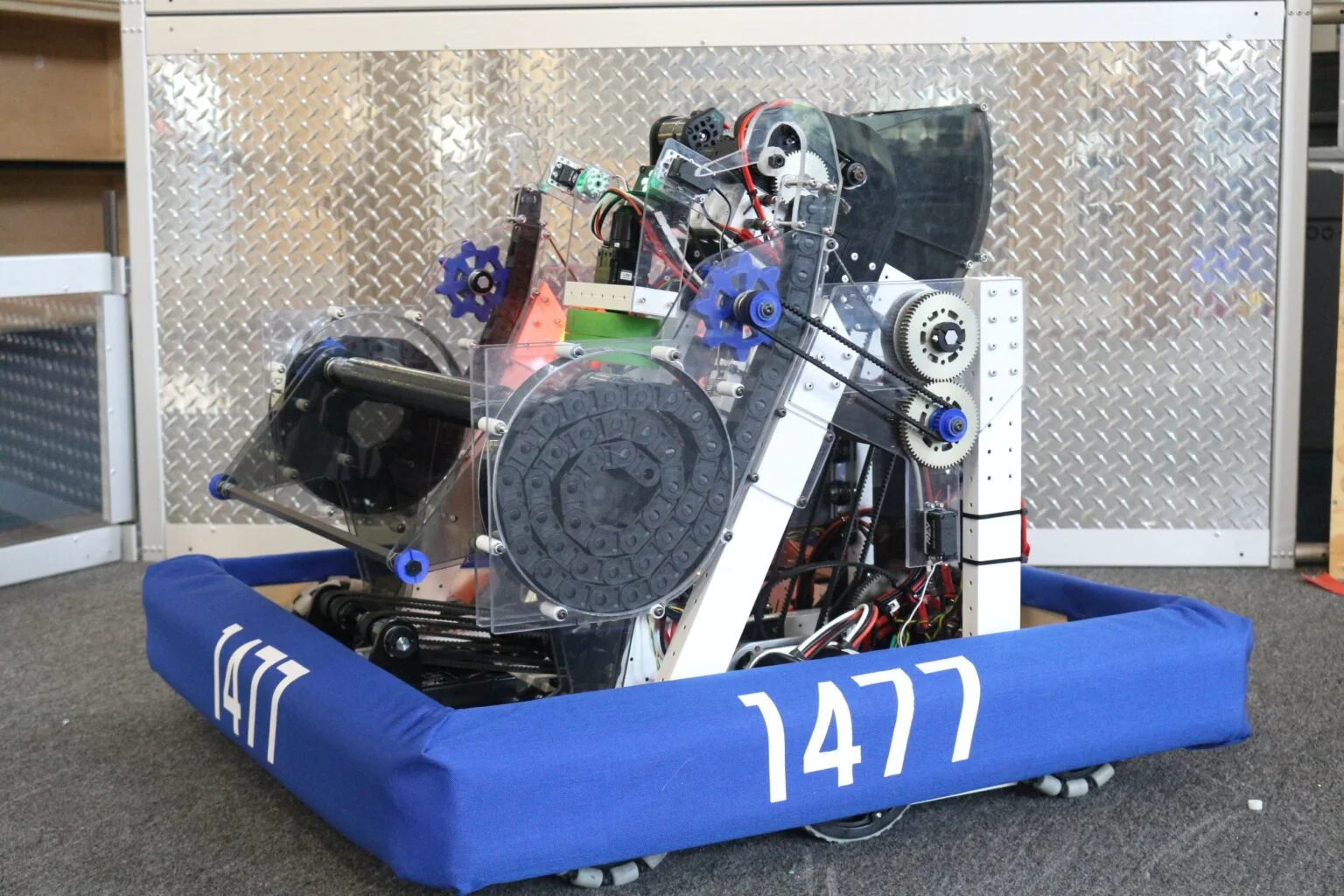 A robot with a metallic frame and transparent panels, housed in a blue padded frame with the number 1477, positioned on a carpeted floor in a workshop setting.