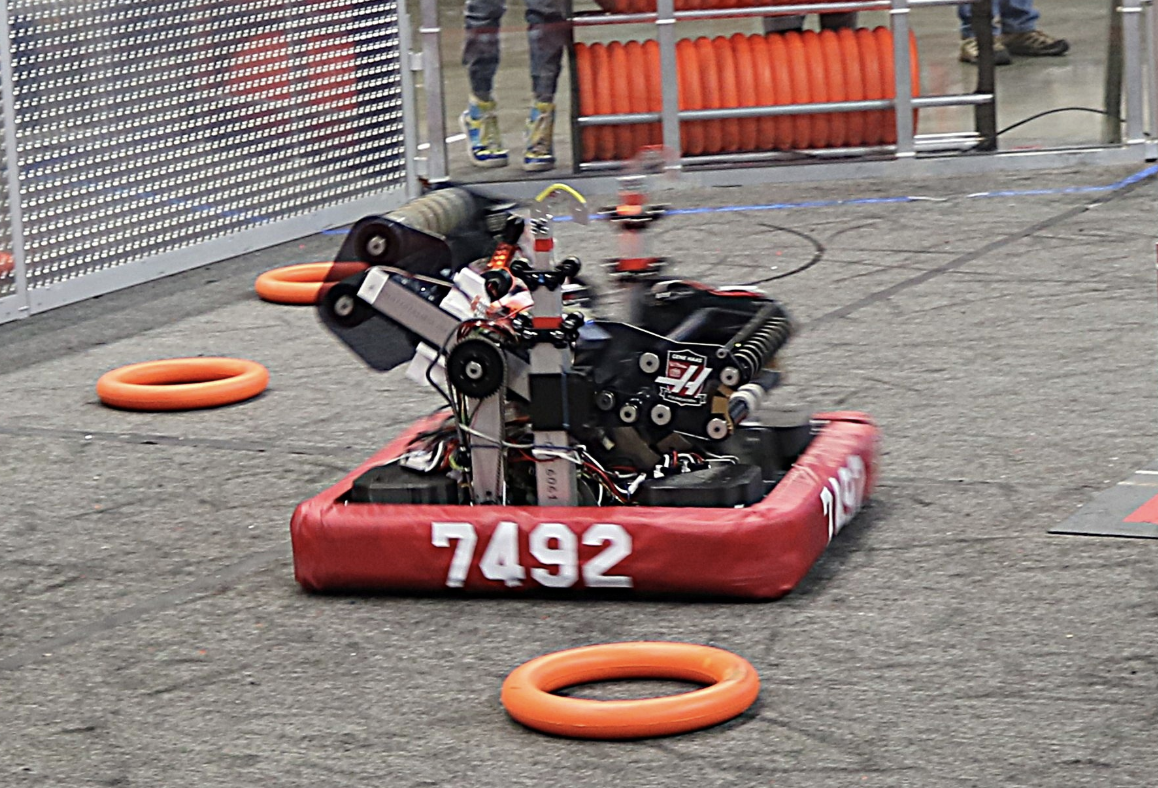 A robot with the number 7492 on it during a robotics competition, surrounded by orange guiding rings on a gray ground, with barriers and spectators in the background.
